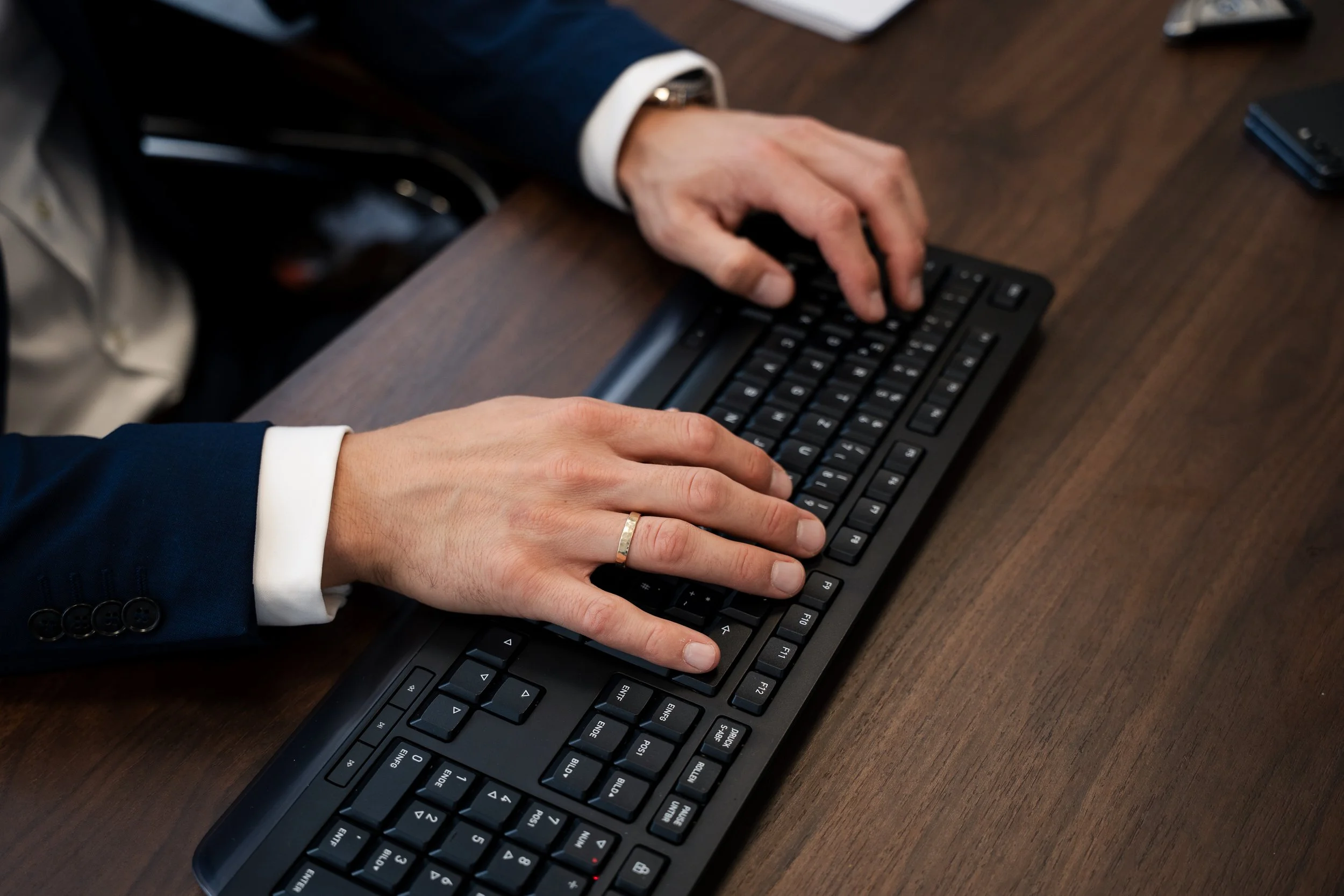 Zwei Hände tippen auf eine schwarze Tastatur auf einem Holzschreibtisch. Eine Person trägt einen blauen Anzug mit weißen Hemdärmeln, die sichtbar sind. Auf dem Tisch sind auch ein Smartphone und einige Papiere zu sehen.