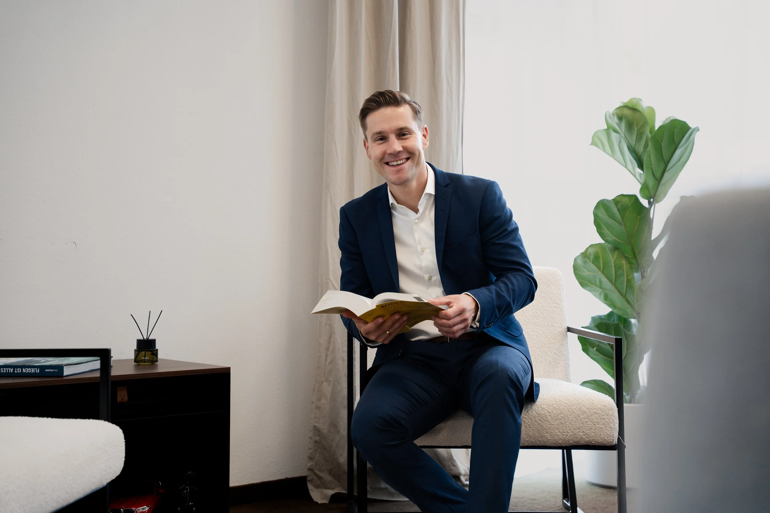Ein Mann in einem blauen Anzug sitzt in einem modernen, hellen Raum, liest ein Buch und lächelt. Im Hintergrund sind weiße Wände, beige Vorhänge, und eine große grüne Pflanze. Auf dem Tisch neben ihm befinden sich Bücher und ein kleiner Diffusor.