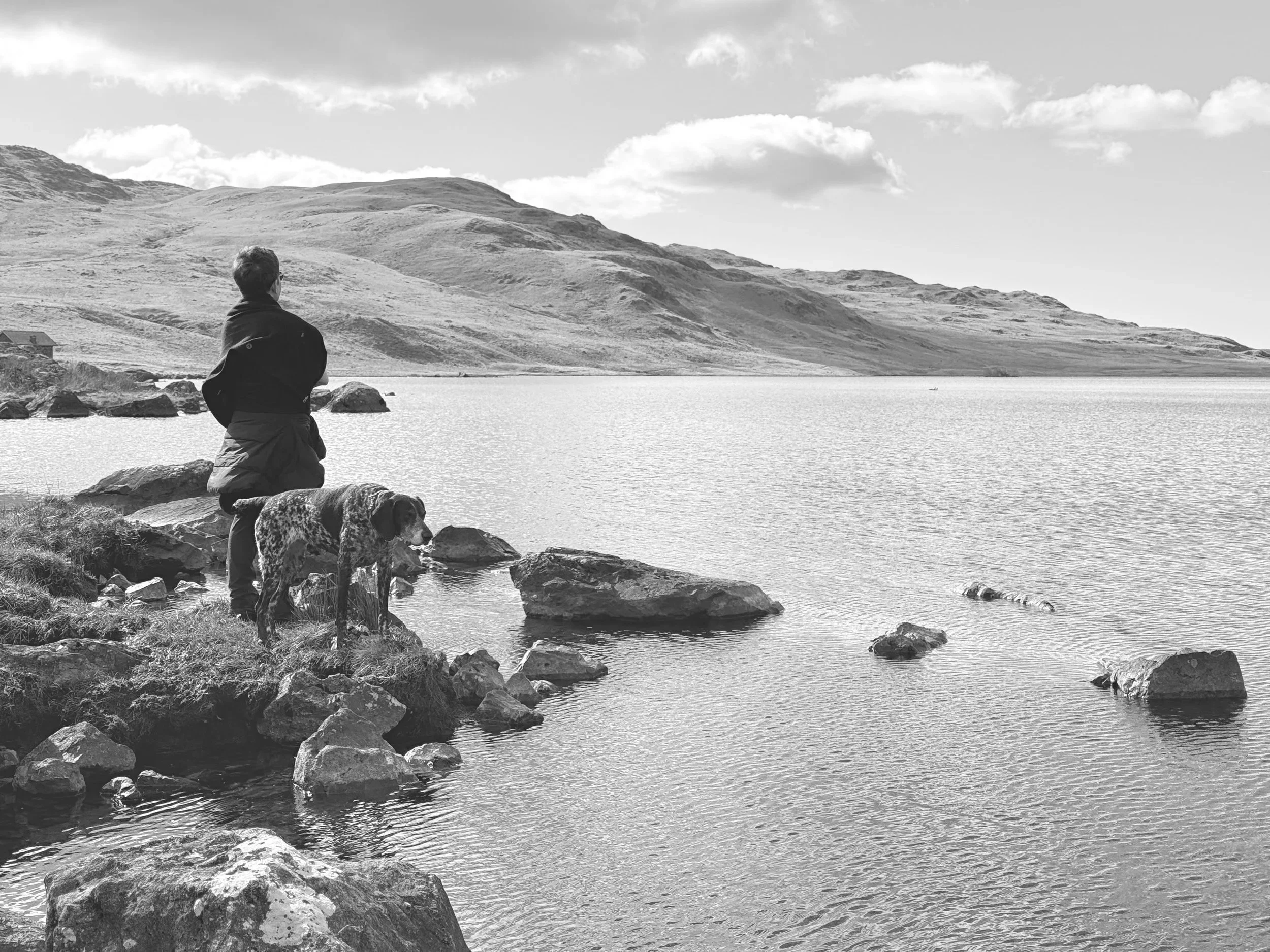Joanna standing on rocks by a lake, accompanied by a German Shorthaired Pointer, with hills in the background and partly cloudy sky.