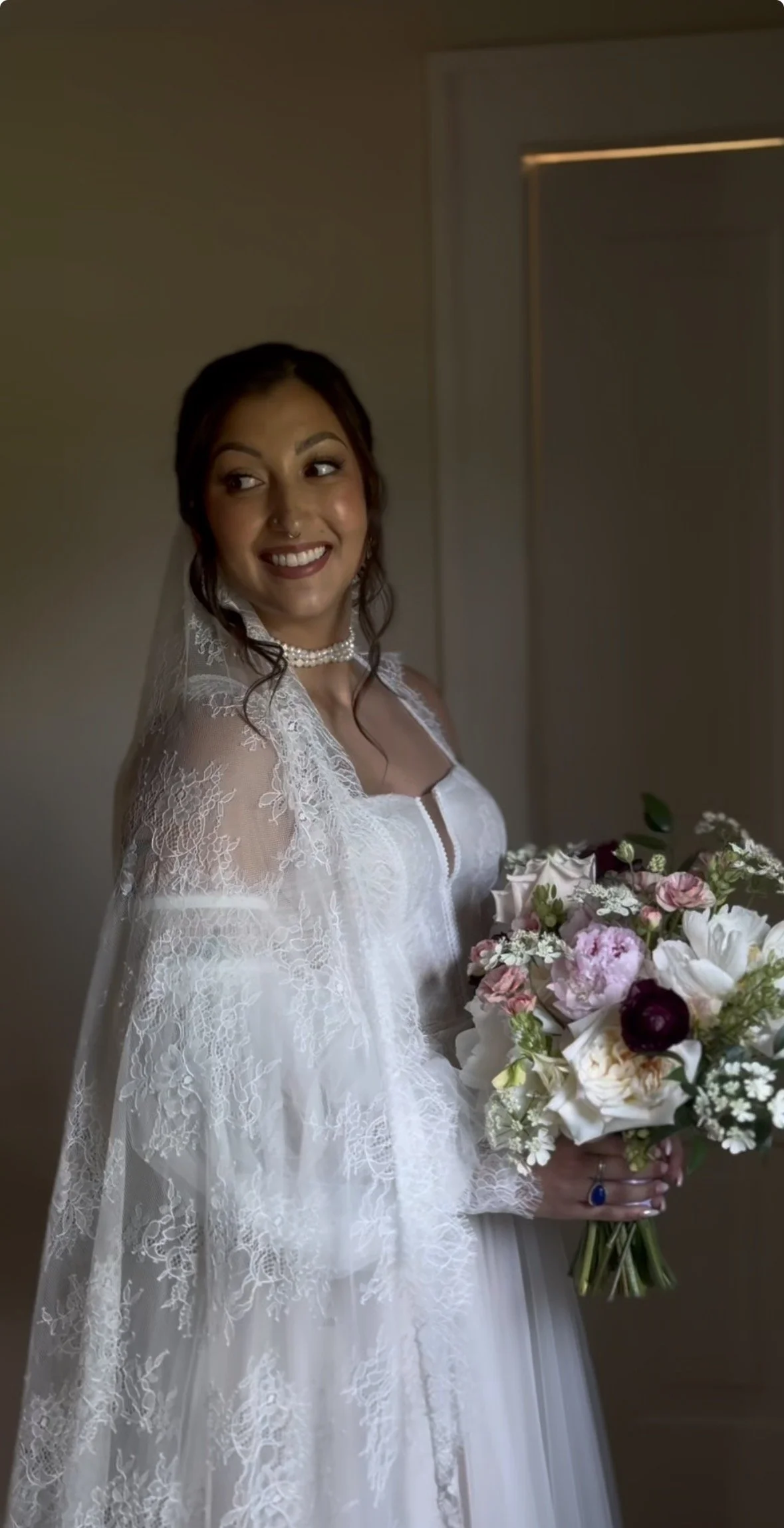 A smiling bride in a white lace wedding gown holding a bouquet of pink, white, and purple flowers, standing indoors near a plain wall and door.