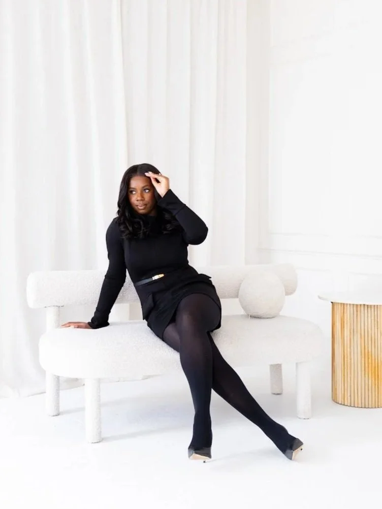 A woman in black seated on a white textured sofa with white curtains in the background, and a small round side table with wooden slats on the right.