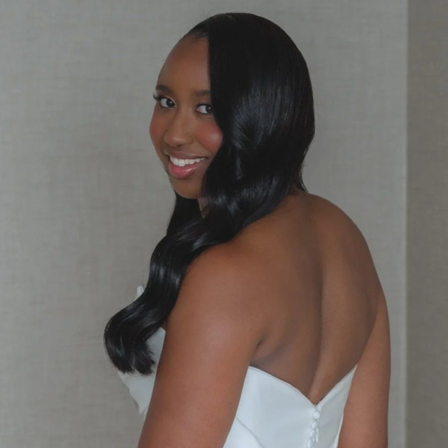 Young woman with long, shiny black hair, smiling, wearing a white strapless dress, standing against a plain neutral background.