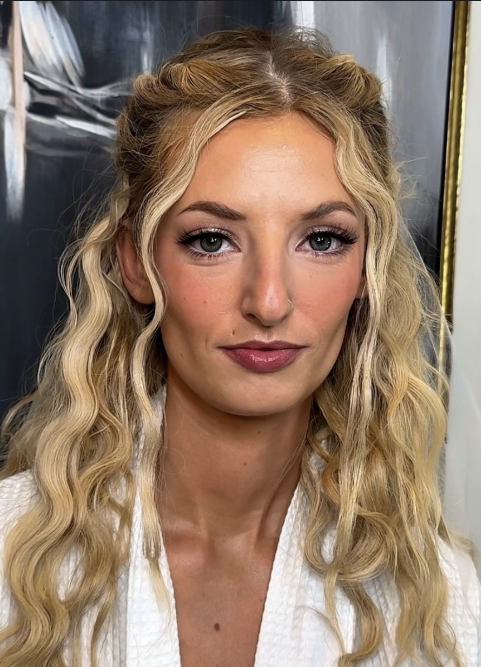 Close-up of a woman with long blonde curly hair, light makeup, and a slight smile, wearing a white garment, against a dark background.