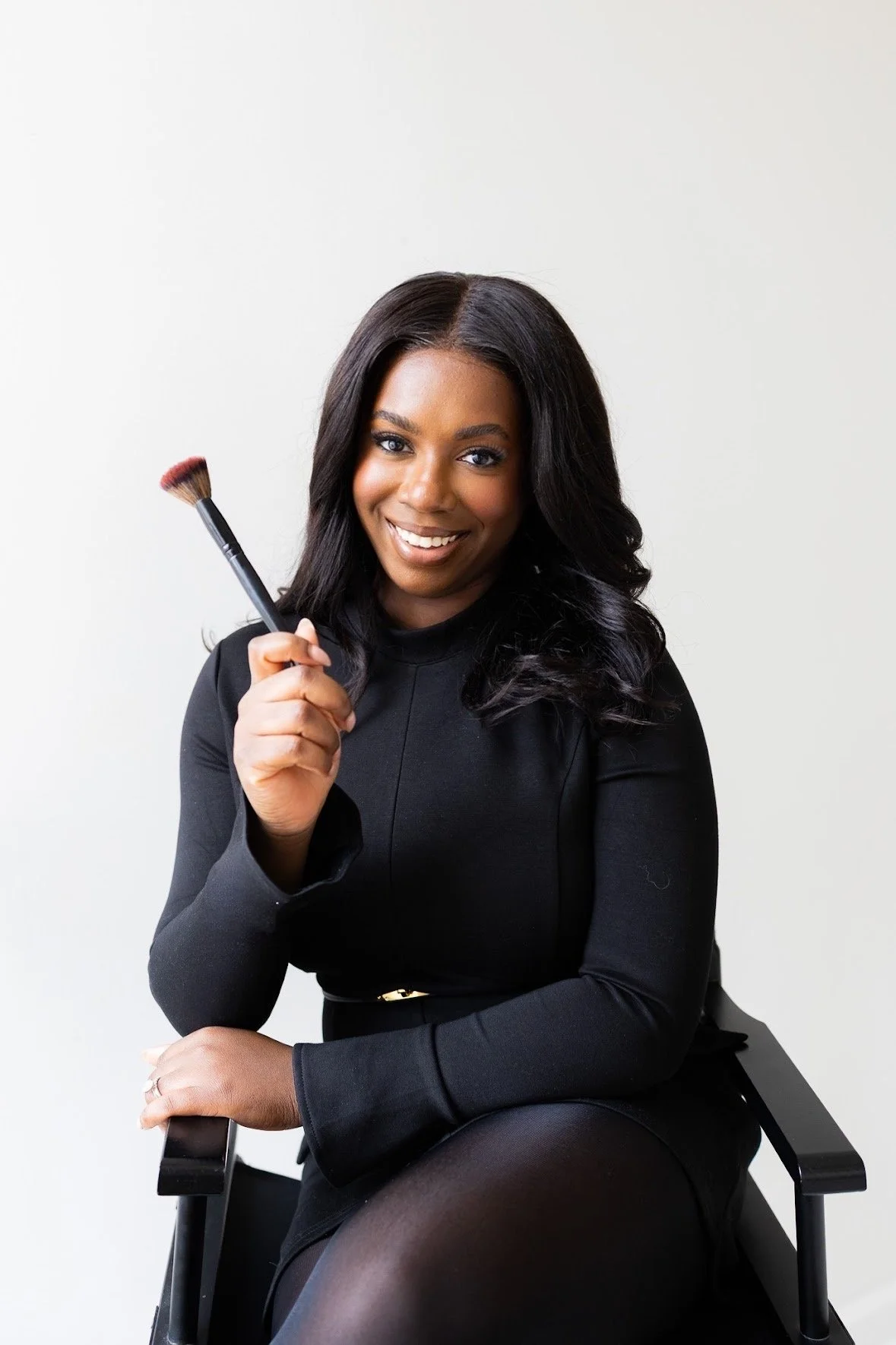 A woman with dark, wavy hair and dark eyes, sitting on a black chair against a plain white background, smiling and holding a makeup brush.