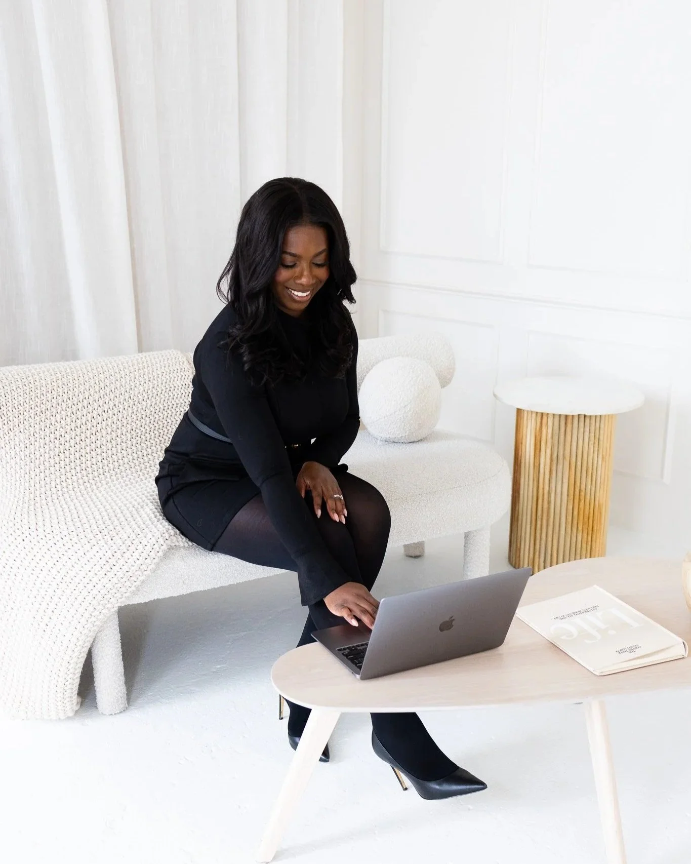 A woman with dark hair smiling while sitting on a white textured chair, looking at her open silver laptop on a small white table in a bright, minimalistic room.