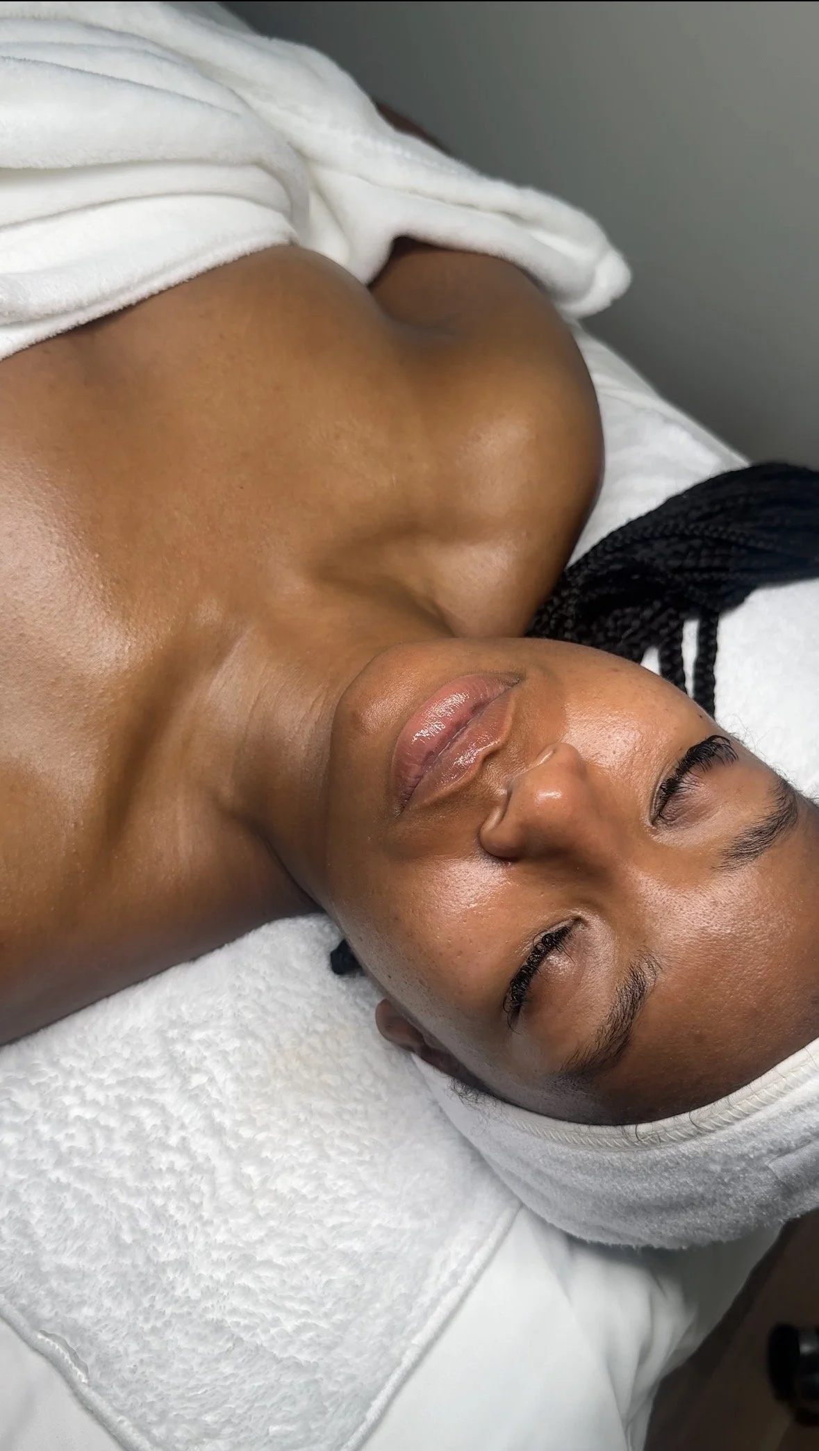 A woman with brown skin and black braided hair lying down with her eyes closed, partially covered by a white towel, on a white towel in a spa or massage setting.