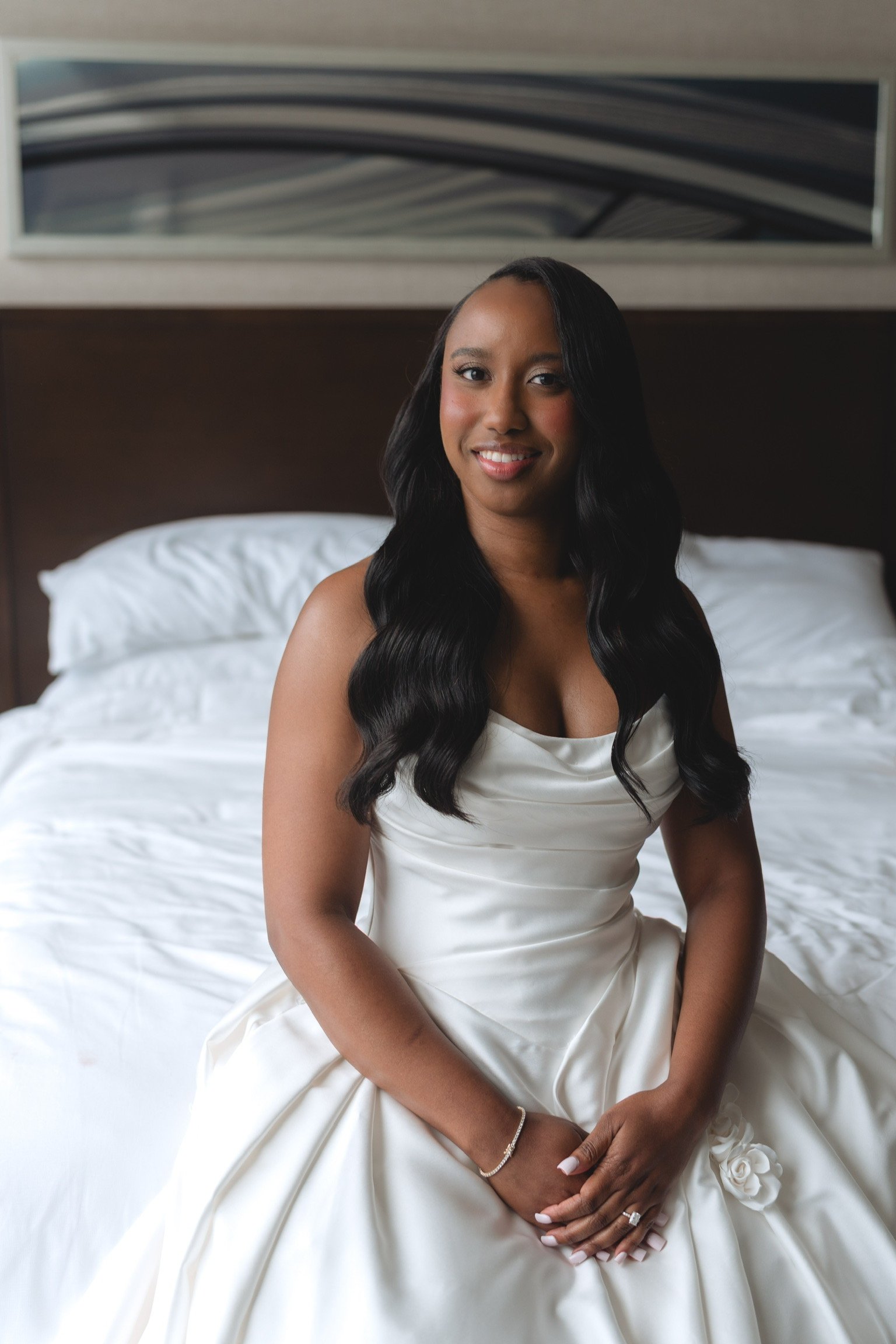 A woman sitting on a bed in a white satin dress in a bedroom, smiling at the camera.