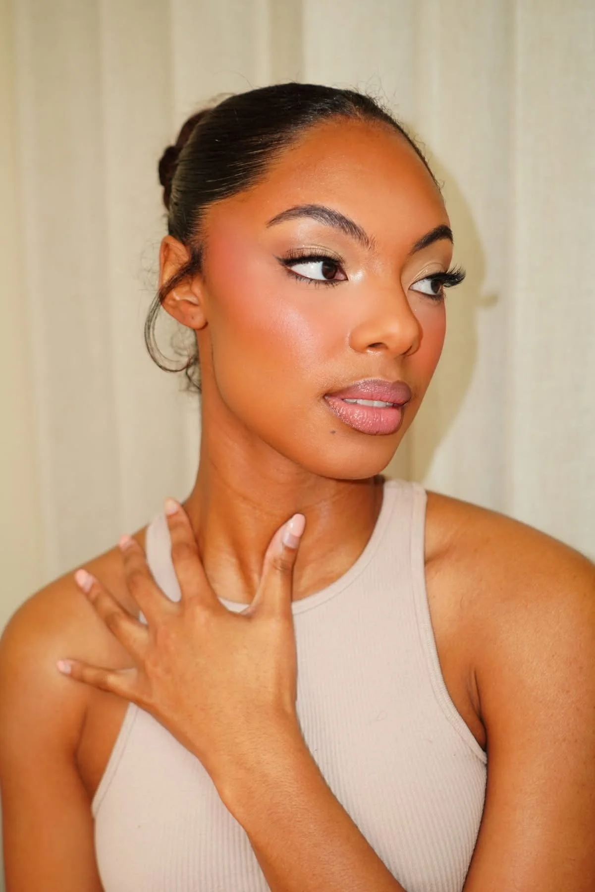 Close-up of a woman with smooth skin, styled hair, wearing makeup, and a light-colored sleeveless top, touching her neck with one hand, standing against a plain background.