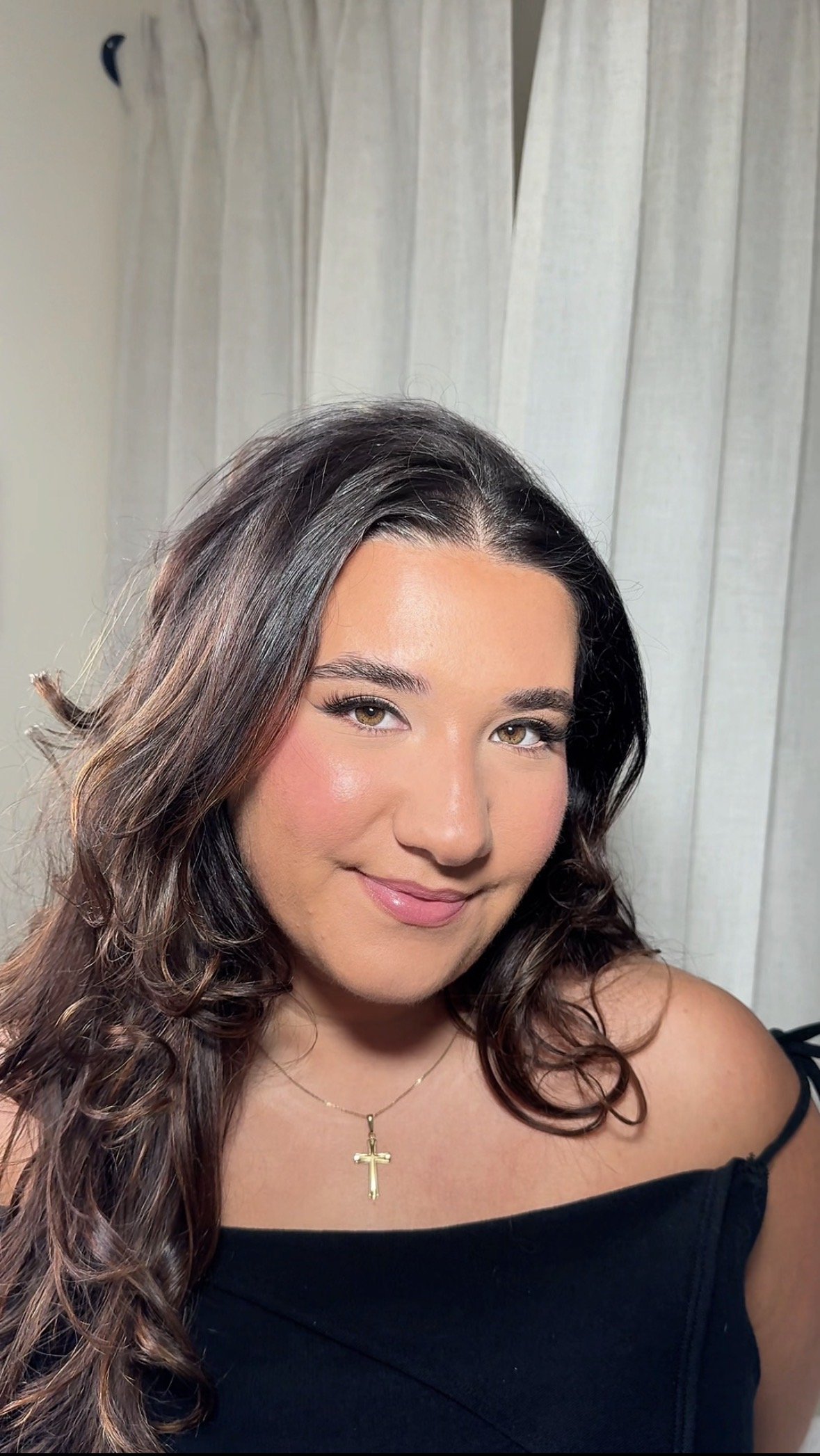 A woman with long, wavy brown hair with highlights, smiling, wearing a black off-the-shoulder top and a gold cross necklace. In the background, there are white curtains.