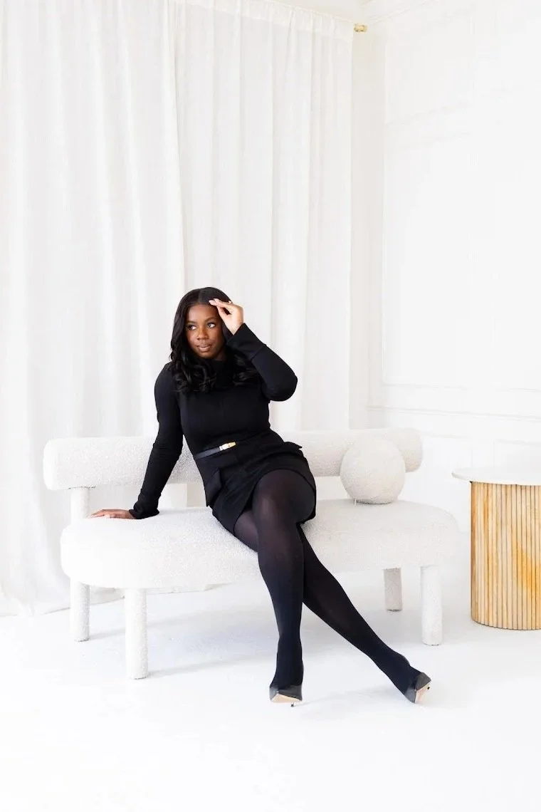 A woman with long dark hair wearing a black dress, black tights, and high heels sitting on a white modern sofa in a bright room with white curtains and walls.