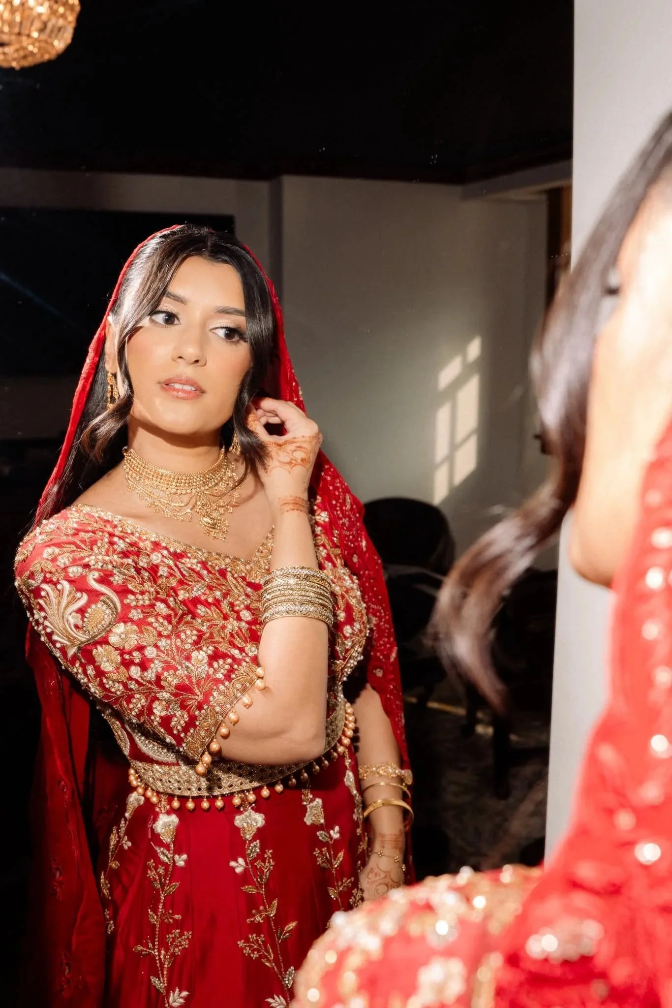A woman dressed in a detailed red and gold traditional South Asian bridal outfit, wearing gold jewelry and mehndi on her hands, looking into a mirror during her wedding preparations.
