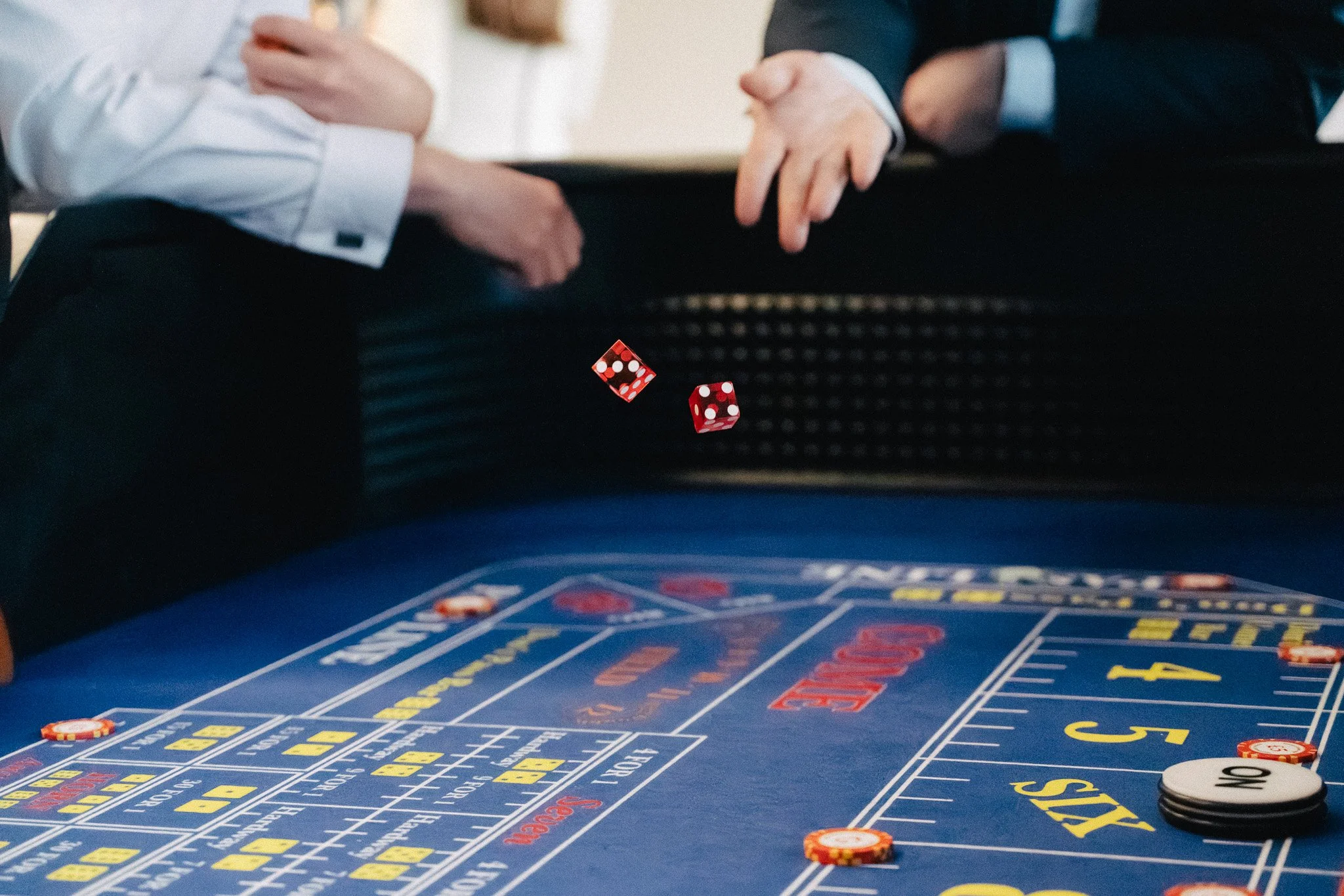 A man in a tuxedo playing roulette at a casino table, placing chips on the green felt surface with other players around.