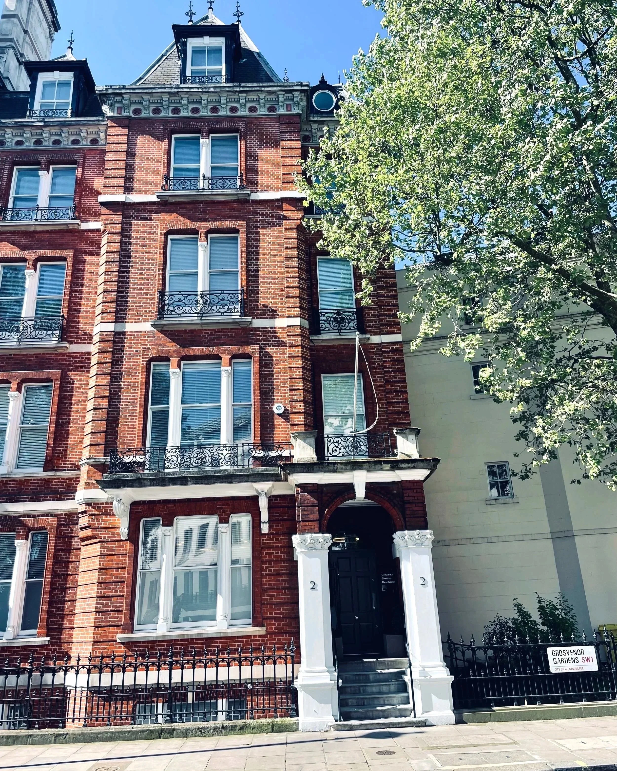 A tall red brick Victorian-style apartment building with ornate iron balconies and white accents, surrounded by a black iron fence and a green tree in front. Nataliya Robinson Clinic