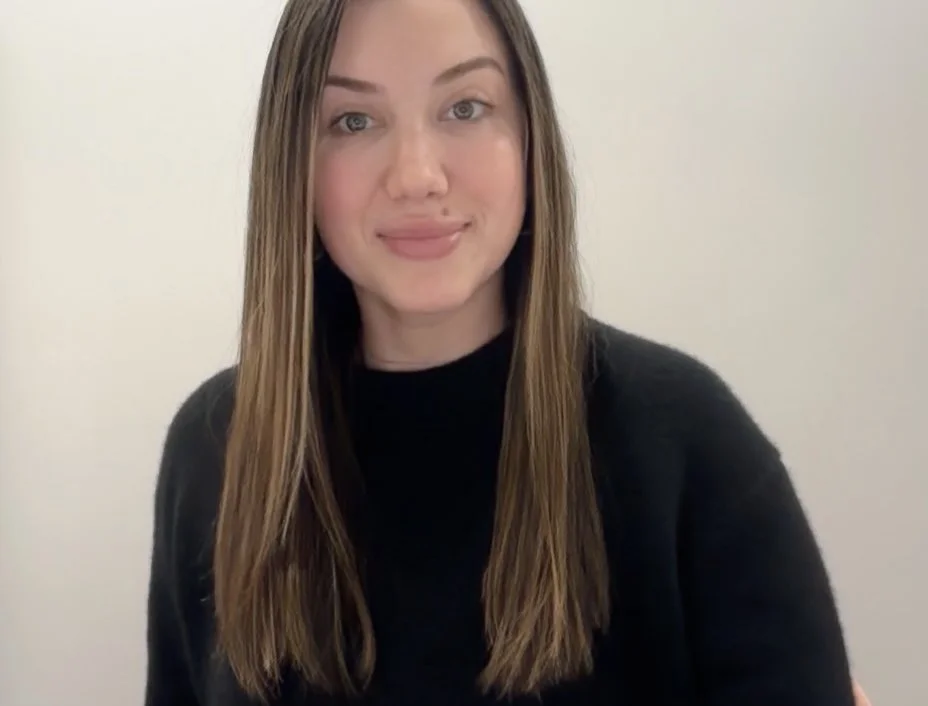 A young woman with long brown hair and a black top smiling at the camera against a plain white background.