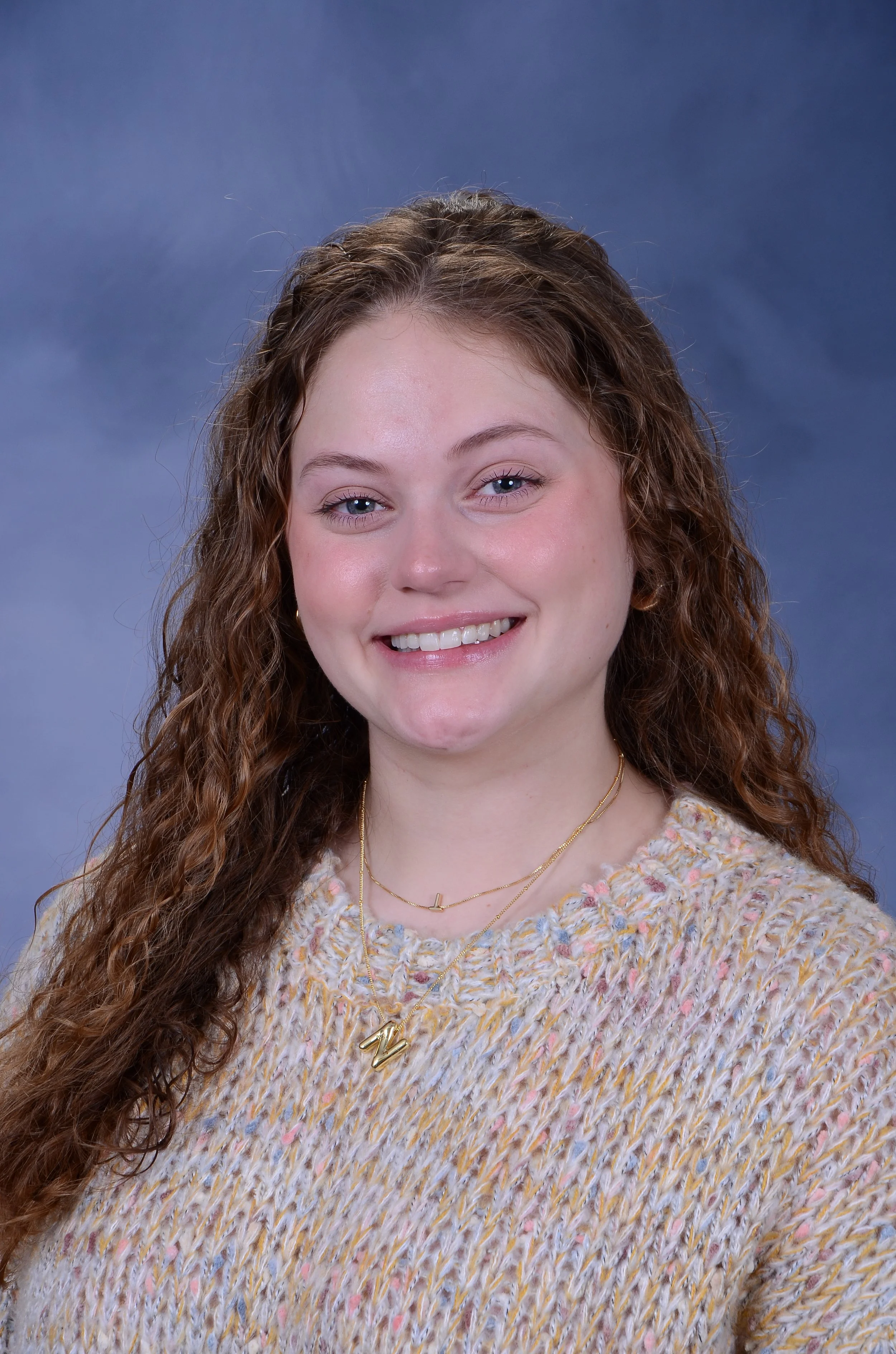 A young woman with long, curly red hair, smiling, wearing a multicolored knitted sweater and layered gold necklaces, posed in front of a blue-gray gradient background.