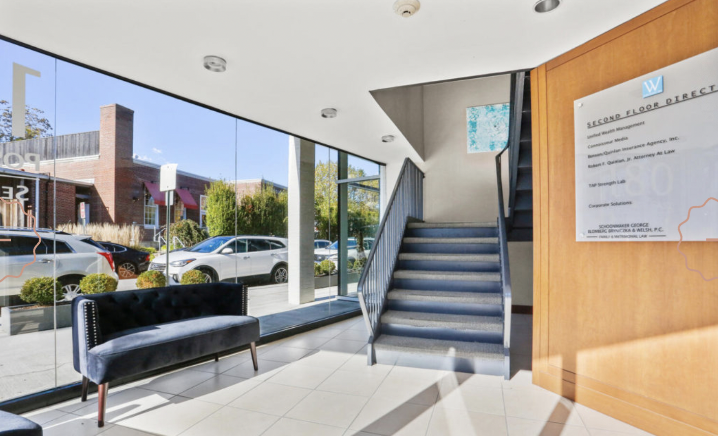 Interior of a building lobby with large glass windows showing parked cars outside, a blue sofa, tiled flooring, a staircase with metal railings, and a wooden wall with a directory sign.