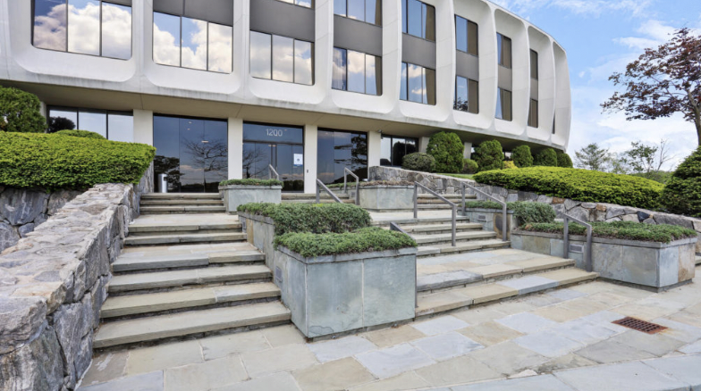 Modern office building entrance with stairs, handrails, and landscaped bushes.