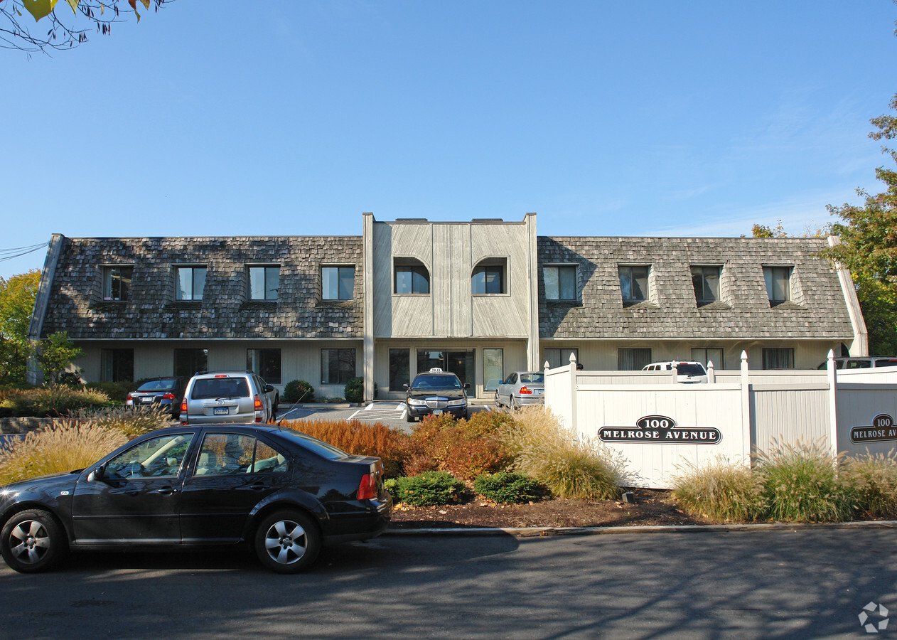 A two-story building with a unique roof design, parking lot with several cars, white fence with a sign reading '100 Melrose Avenue', and landscaped plants in front.