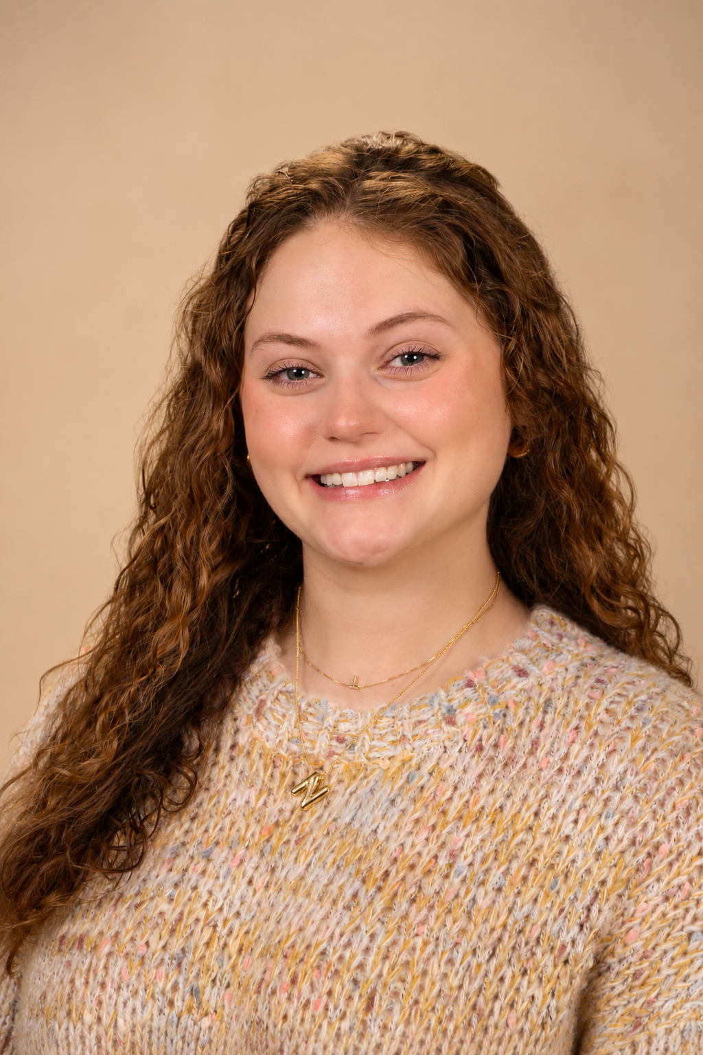 A young woman with long, curly brown hair smiling at the camera, wearing a multicolored knit sweater and layered gold necklaces with pendant charms, posing against a plain beige background.