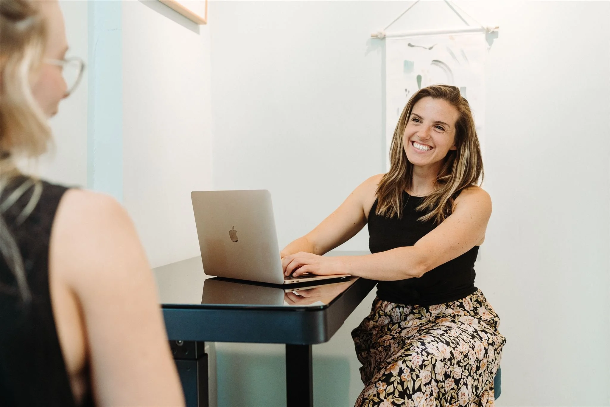 A woman with shoulder-length brown hair, wearing a black sleeveless top and floral skirt, smiling while sitting at a desk with a silver laptop, engaging in conversation with another woman whose shoulder and ear are visible in the foreground.