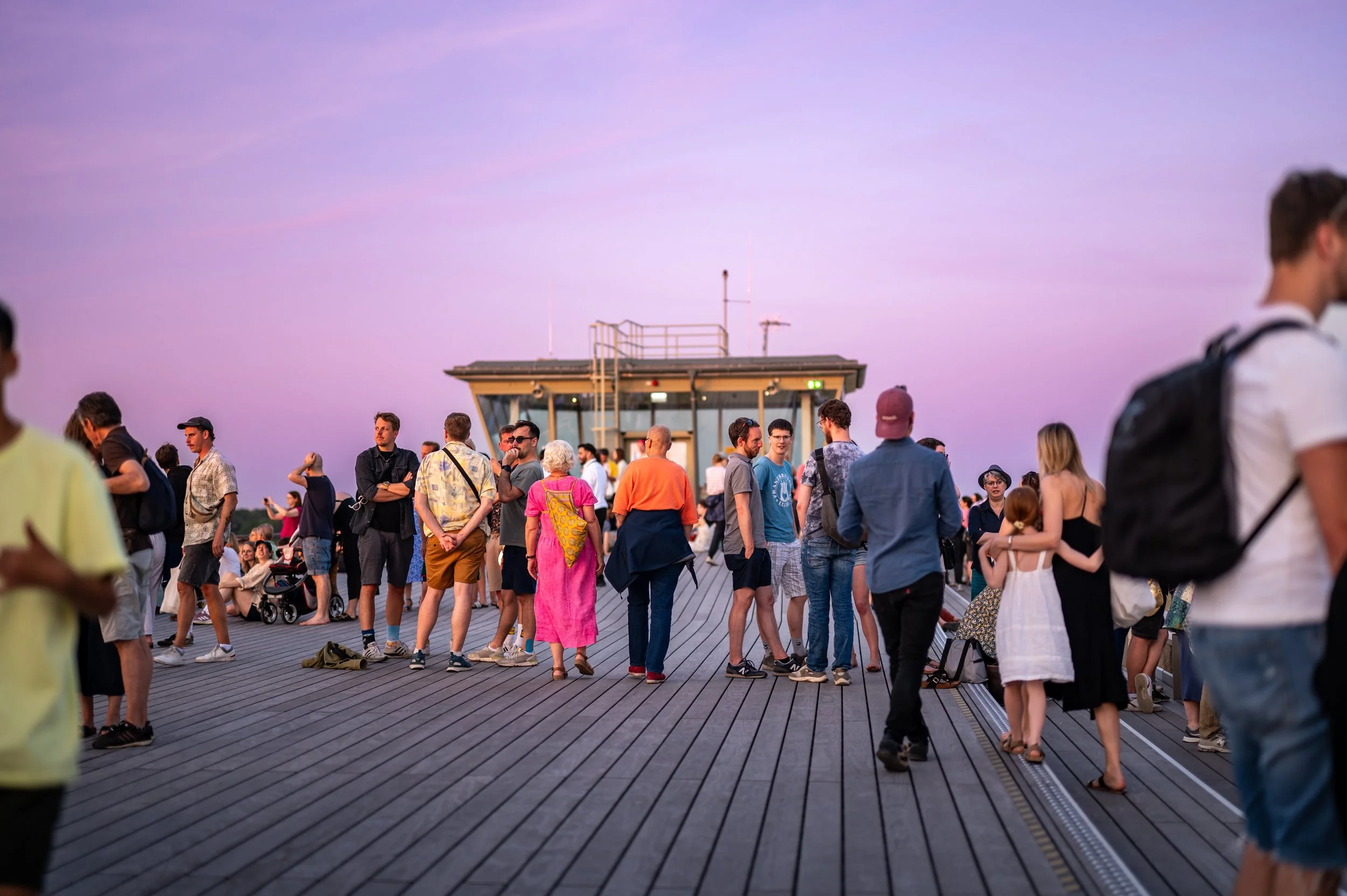 People gathered on a wooden pier during sunset, some talking and others taking photos, with a small building or observation station in the background and a purple sky overhead.