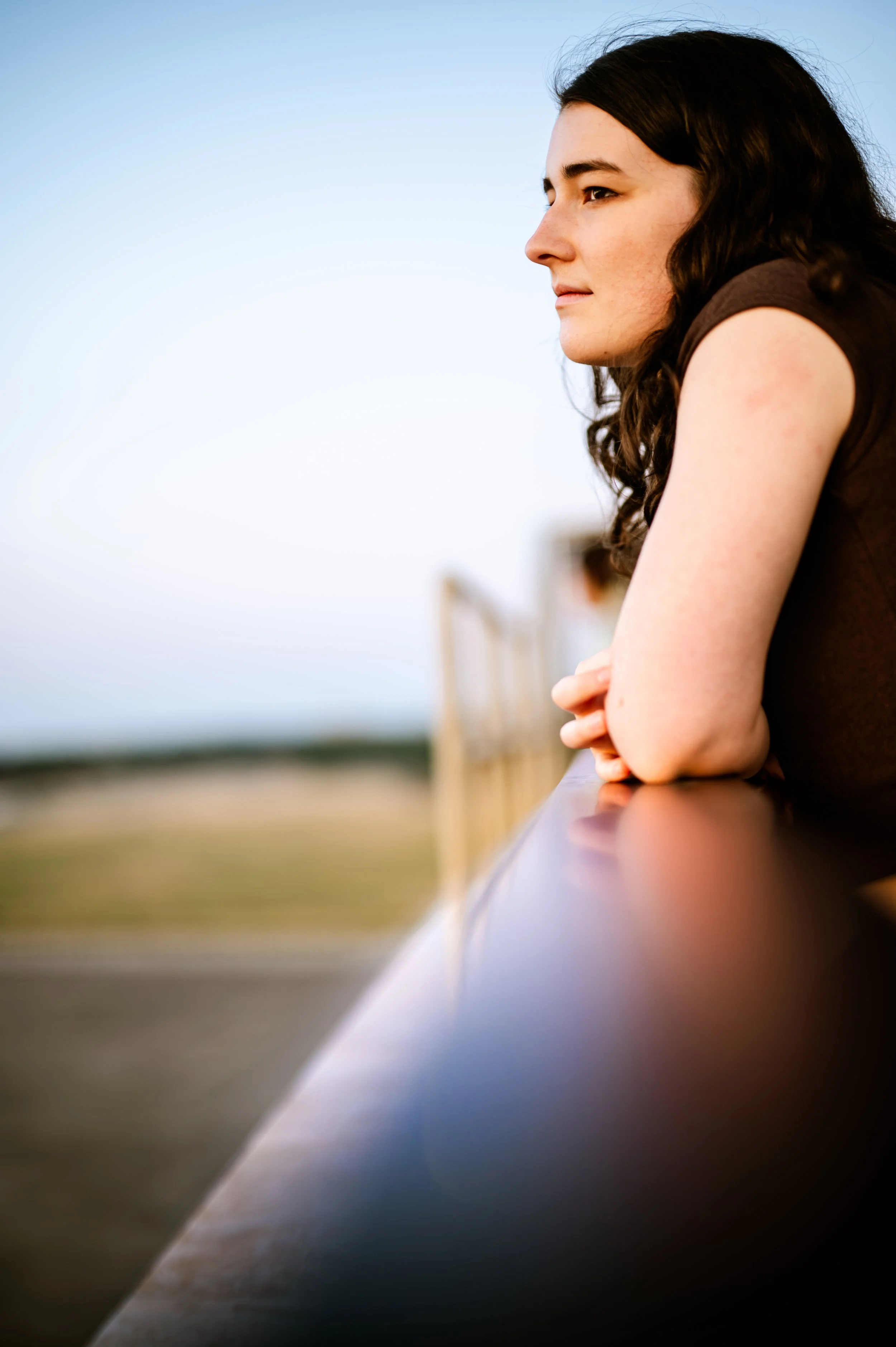 A woman with dark, curly hair leaning on a railing outdoors during the daytime, gazing thoughtfully into the distance.