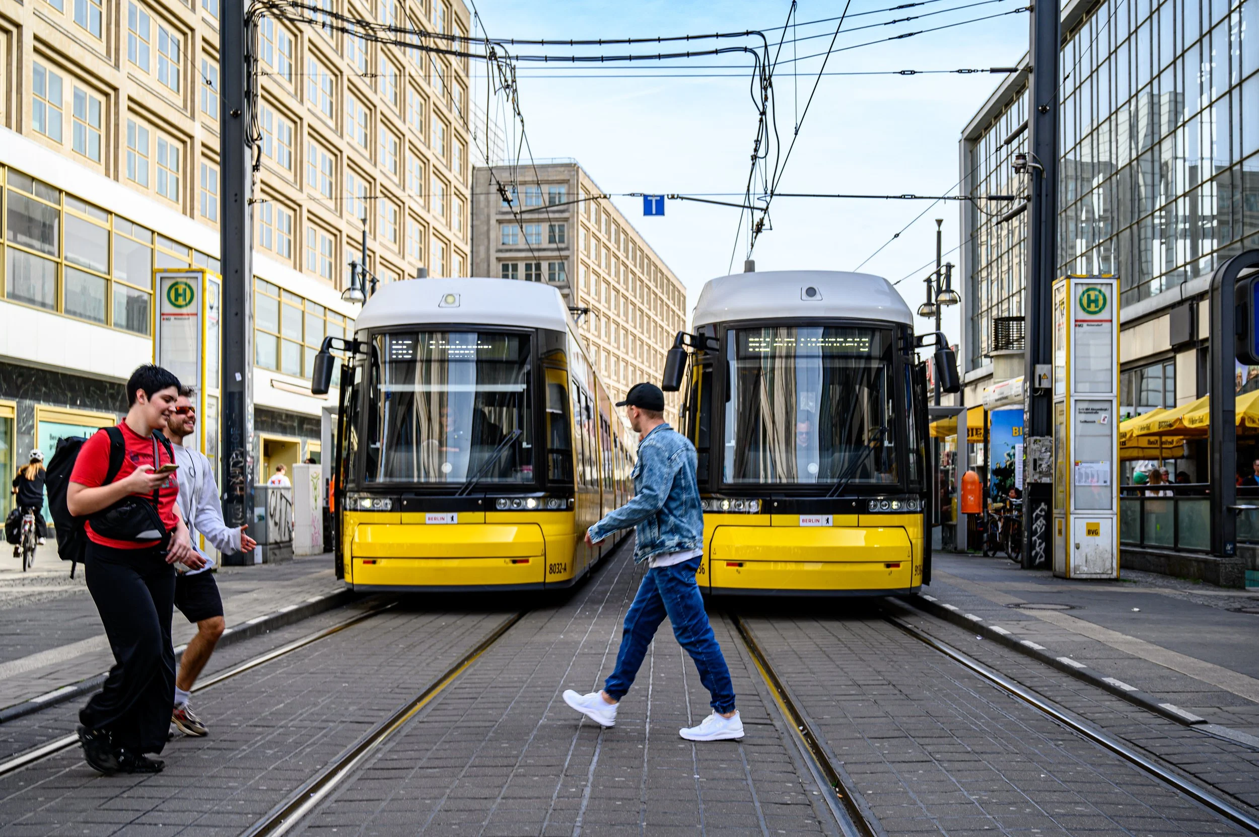 Two yellow trams on city tracks with pedestrians crossing in front of them, in an urban area with tall buildings and overhead power lines.