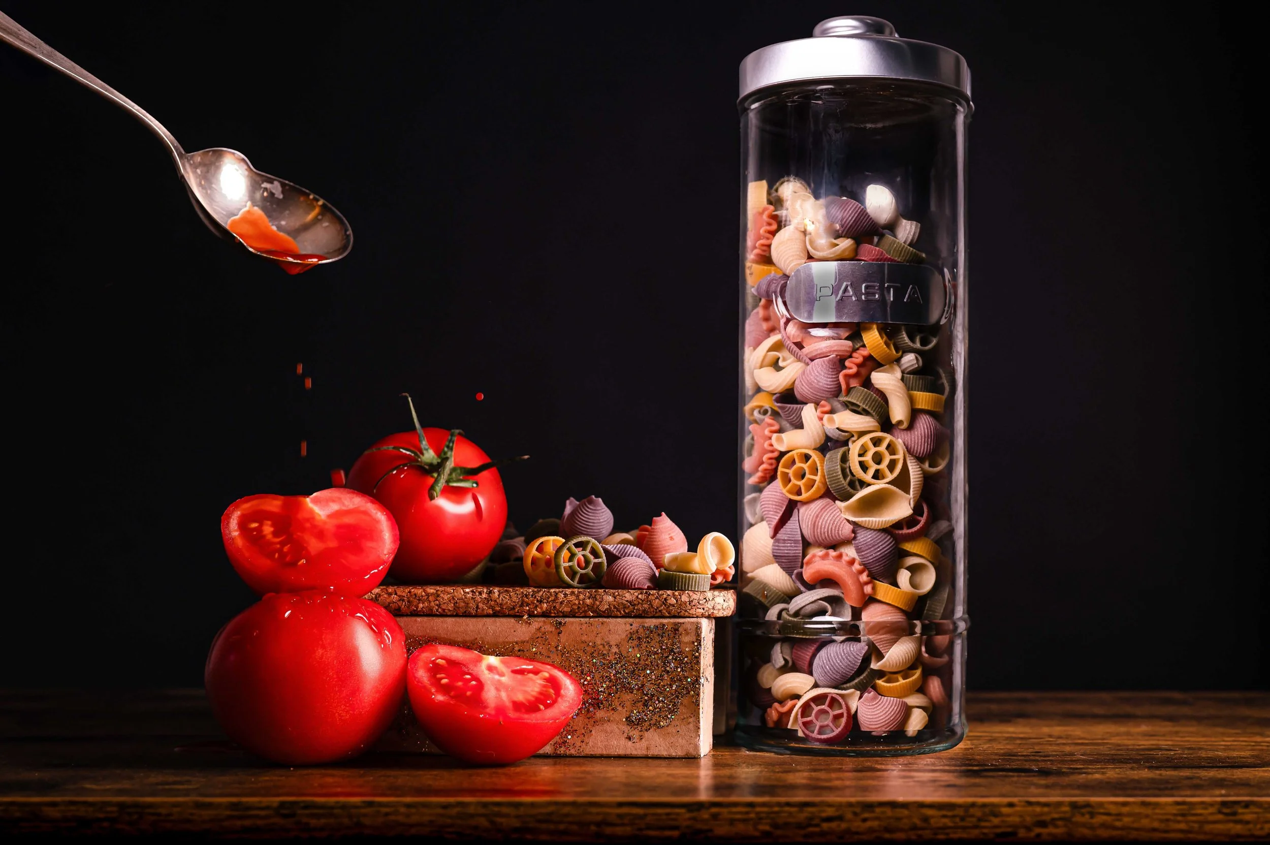 A glass jar filled with colorful pasta on a wooden surface, with fresh tomatoes and a spoonful of sauce nearby against a dark background.