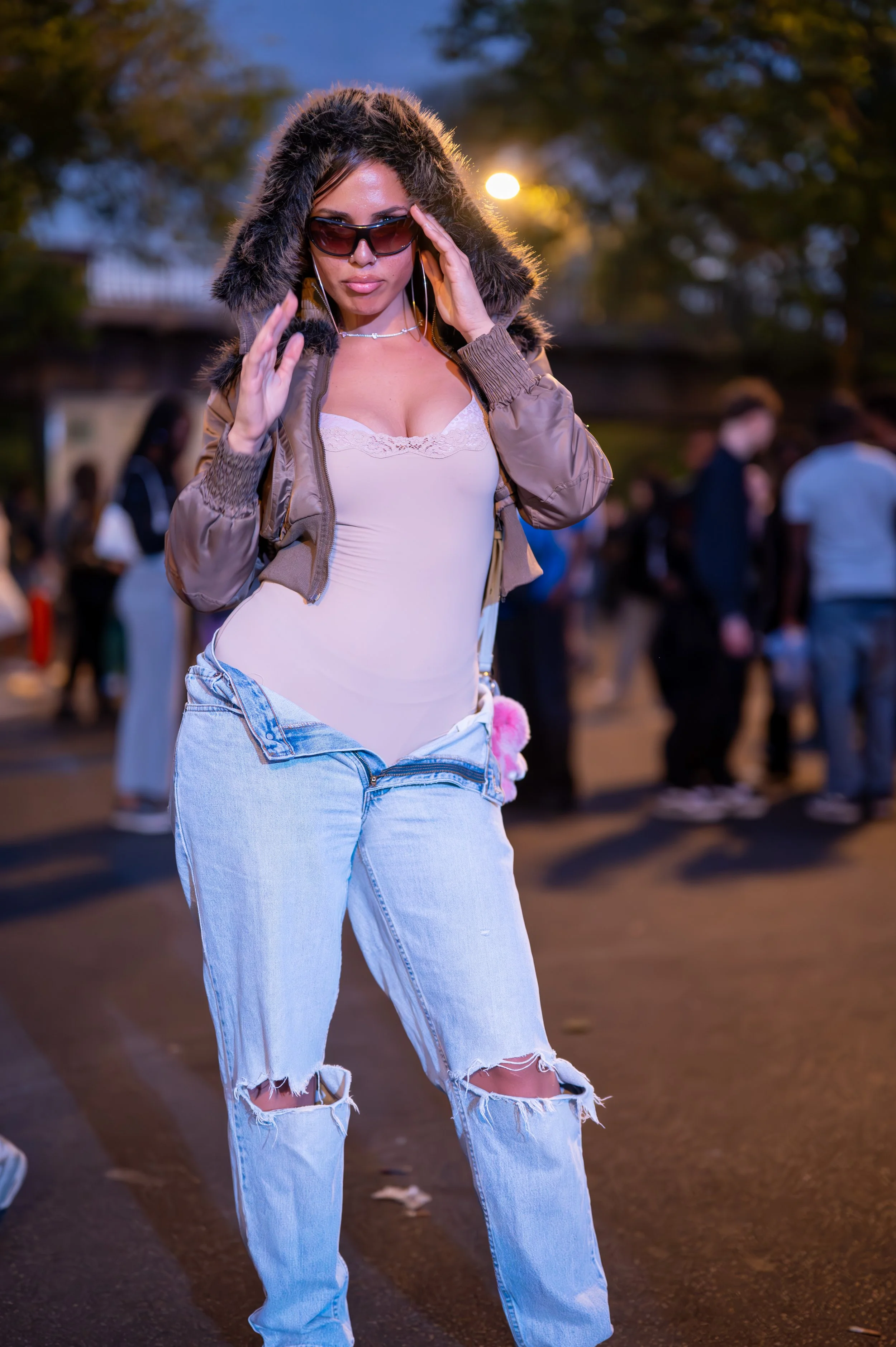 A woman wearing sunglasses, a fur-lined hooded jacket, a white top, and ripped jeans standing outdoors during evening hours, with a crowd of people in the background.