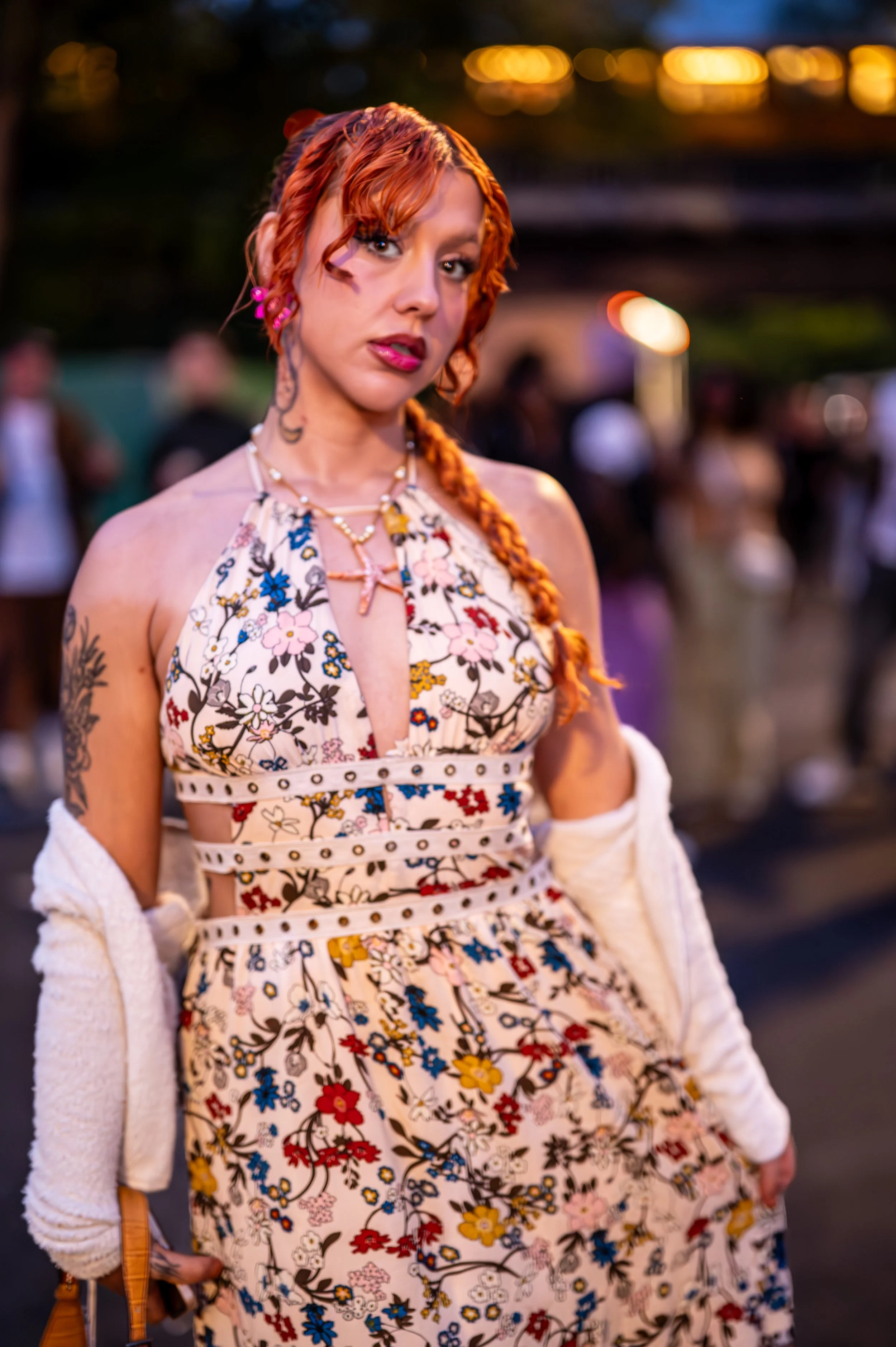 A woman with red hair in a braid, wearing a floral dress with cutouts, standing outdoors at dusk with blurred people in the background.
