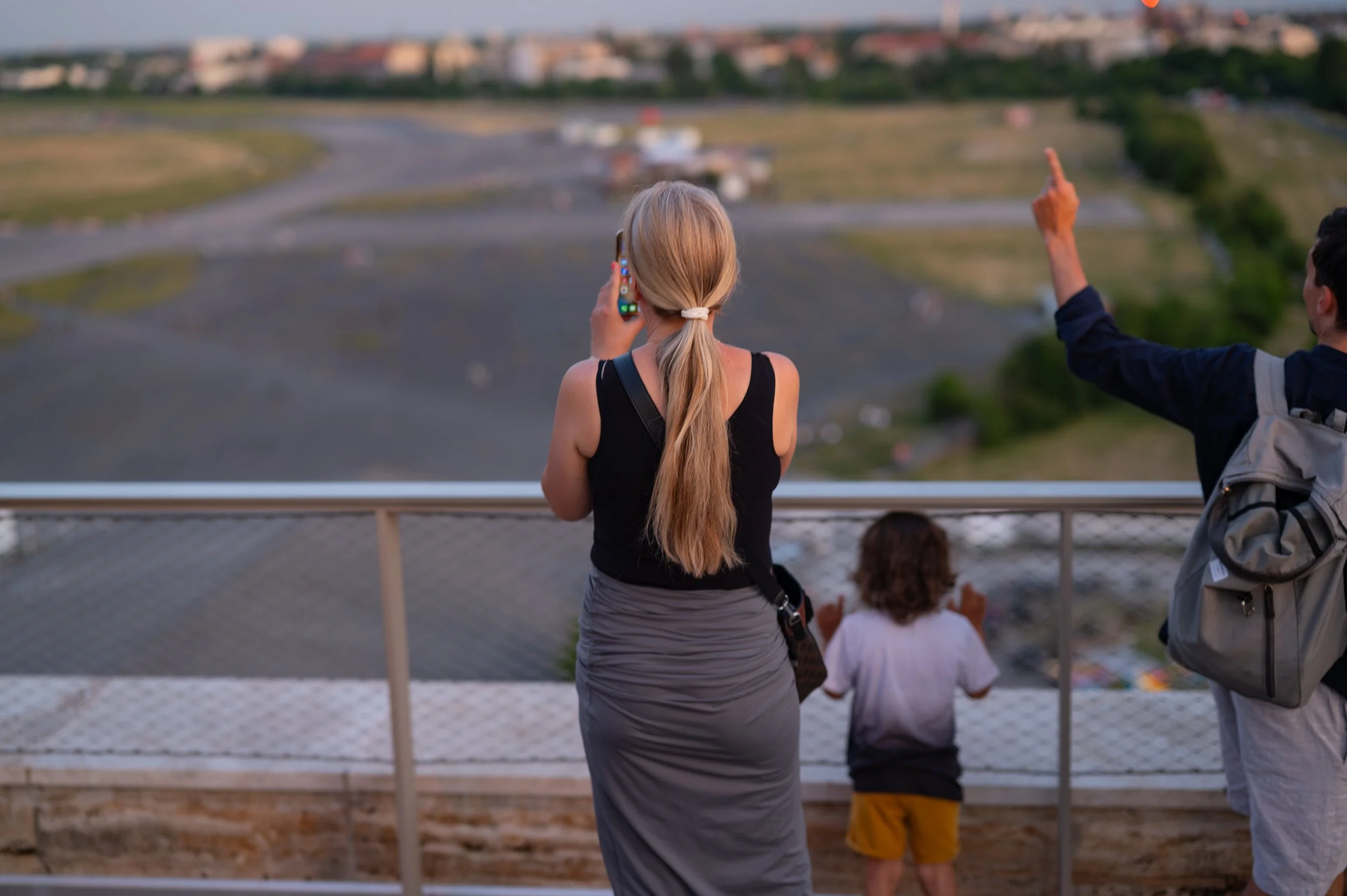 A woman, child, and man observe and take photos of a landscape from an observation deck. The woman is talking on her phone, the child is looking out at the view, and the man is pointing at something in the distance.