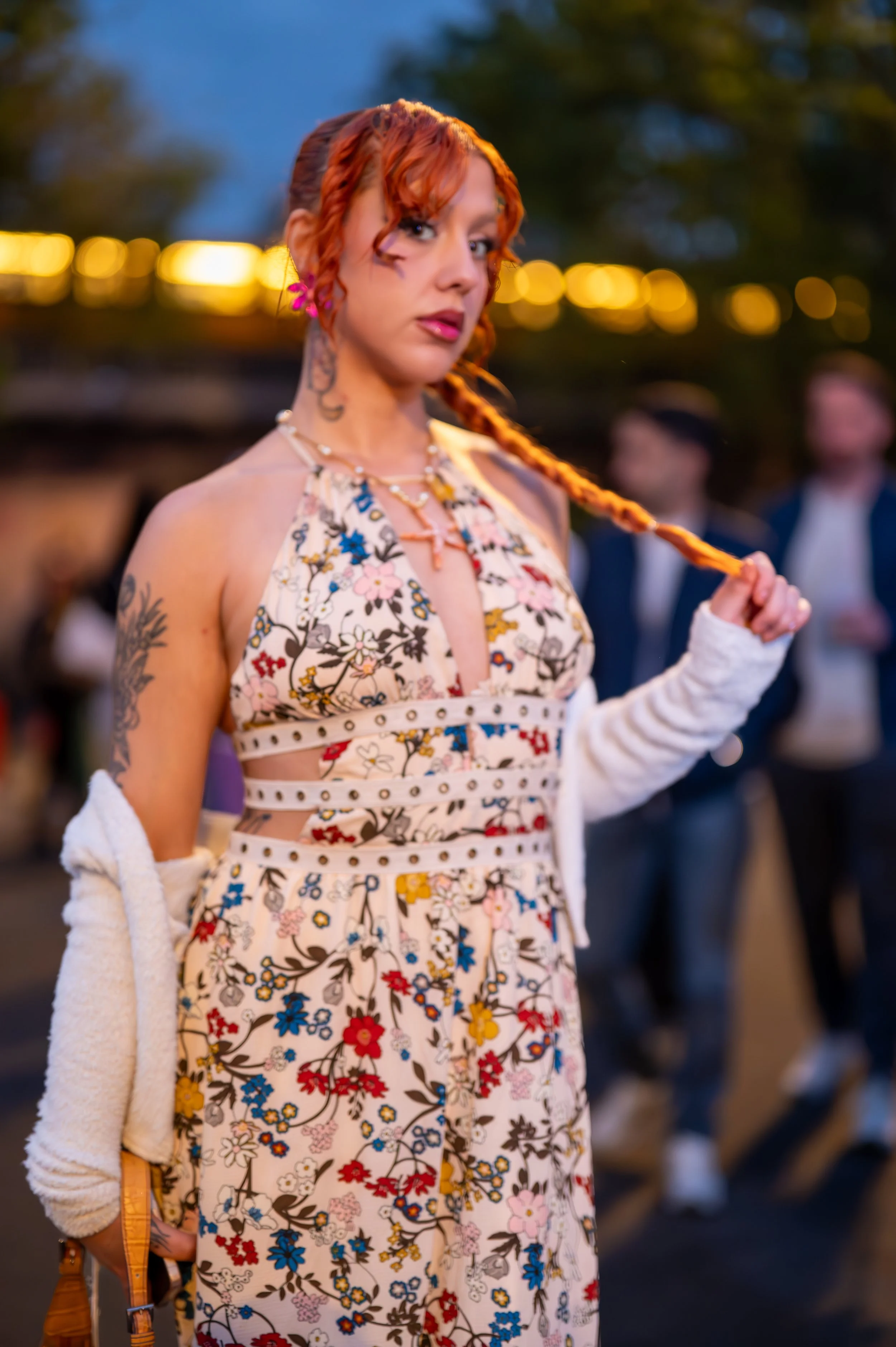 A woman with red curly hair and tattoos wearing a floral dress and white cardigan, holding her braid, standing outdoors during the evening. Background includes blurred people and warm lights.
