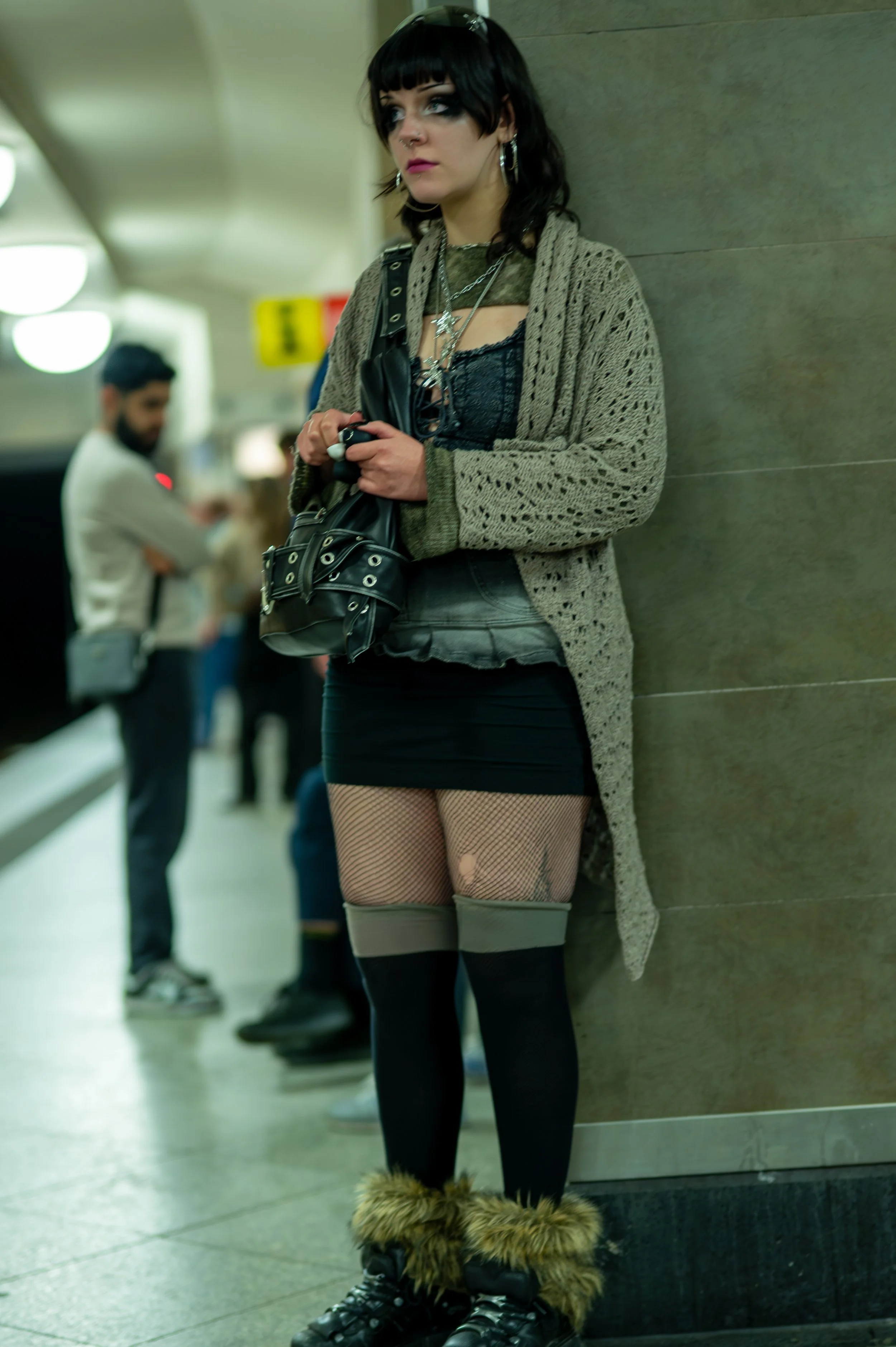 A woman with dark hair and piercings standing in a subway station, dressed in a goth-inspired outfit with fishnet stockings, fur-lined boots, and layered jewelry.