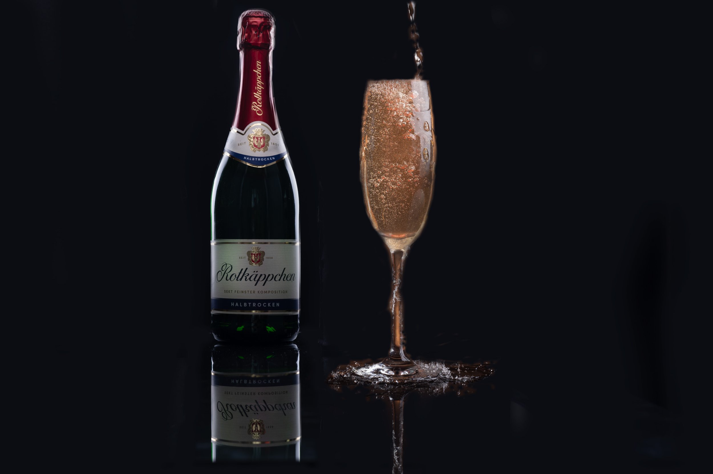 A bottle of Rotkäppchen sparkling wine and a glass of sparkling wine being poured, against a black background with reflections.