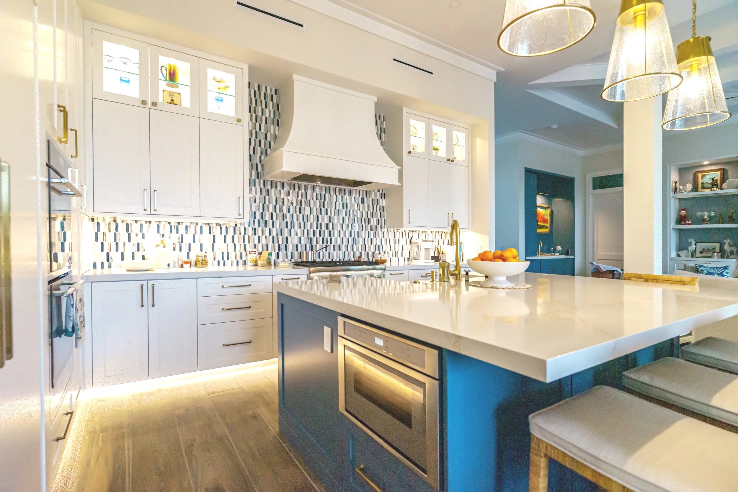 Modern kitchen with white cabinetry, a marble island with blue base, and a patterned tile backsplash with shades of blue, white, and gray; gold accents on fixtures and lighting; a bowl of oranges on the island; open shelving with decorative items; bright and airy atmosphere.