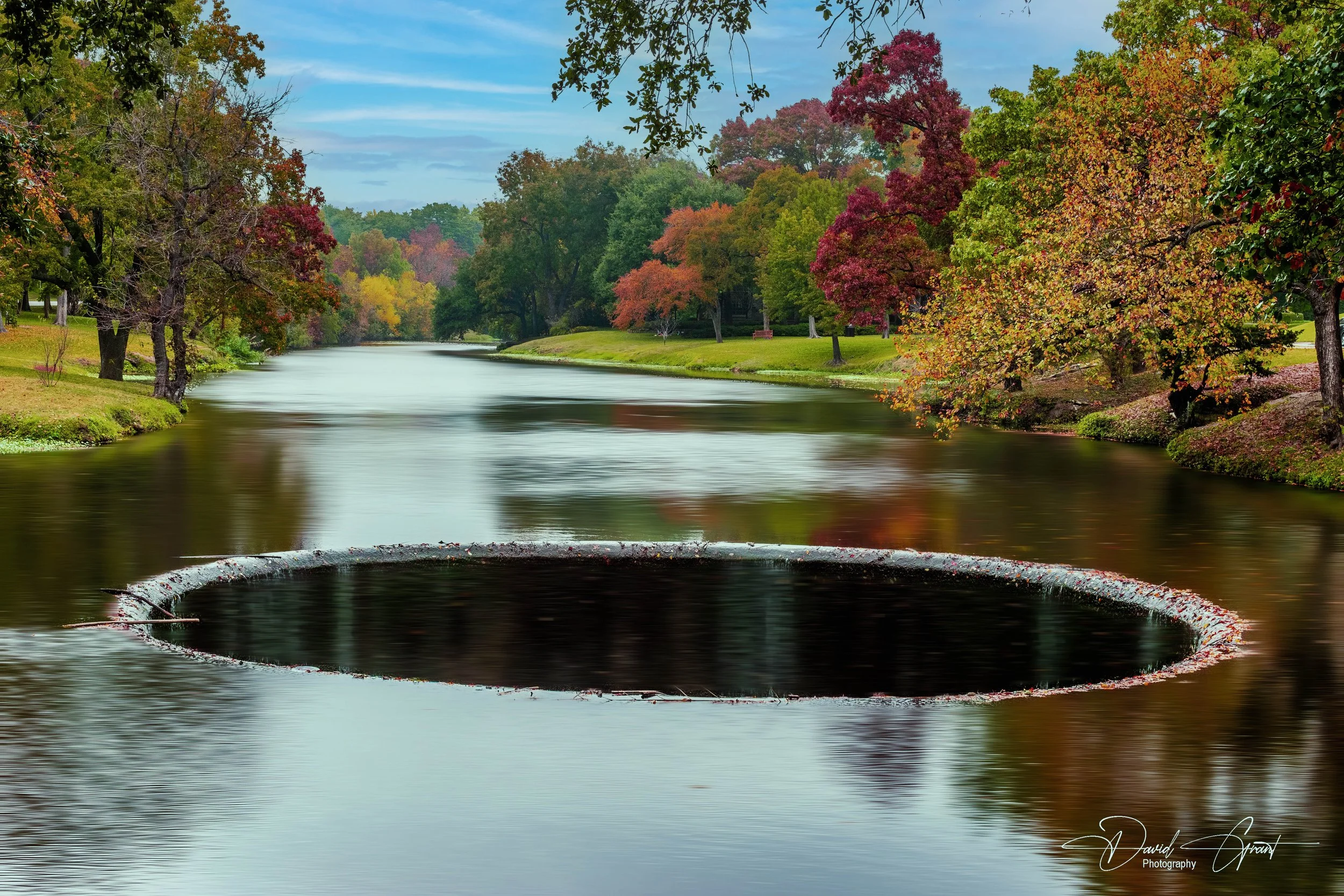 A river running through a park with trees displaying fall colors and an open manhole cover in the water.