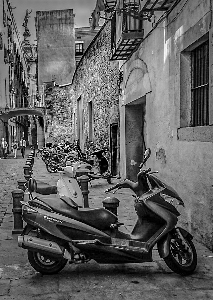 Scooter parked on a narrow cobblestone street with more scooters and pedestrians in the background, historic buildings lining the street, and a stone archway in an urban setting.
