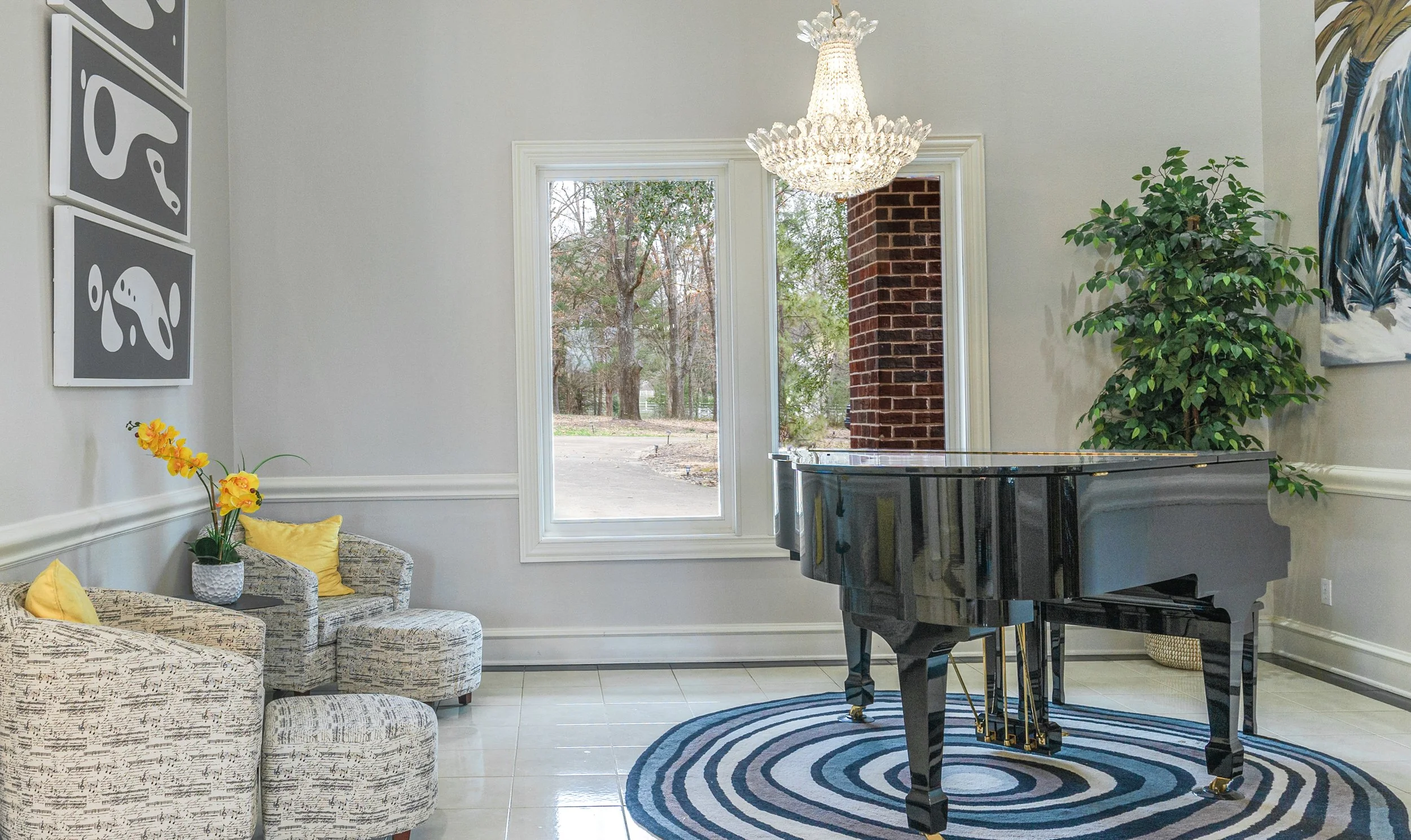 Living room with a grand piano, a large window revealing an outdoor scene, a green potted plant, black and white artwork, a chandelier, and seating with yellow pillows.