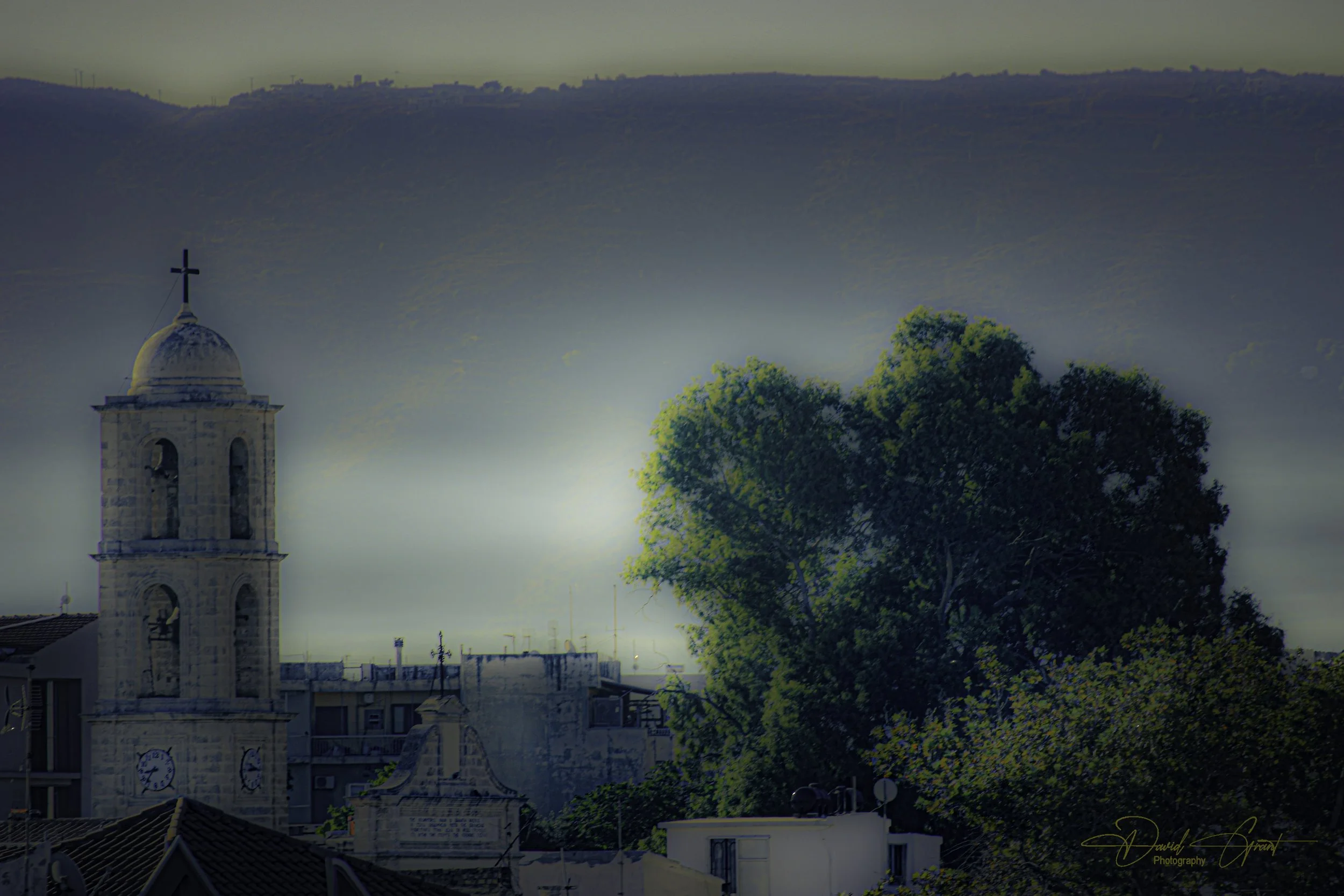 A church tower with a cross at the top, a large green tree, and a cityscape with buildings and rooftops under a cloudy sky.