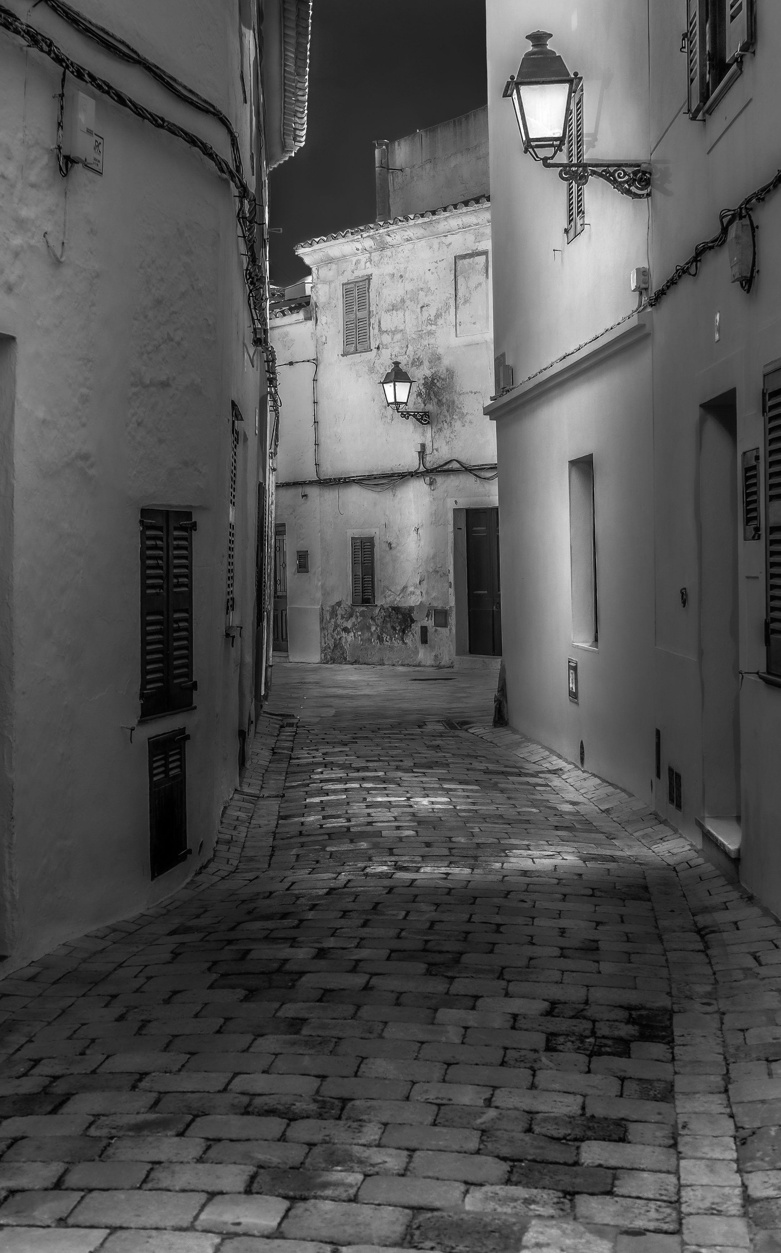 A narrow, winding cobblestone street in a European town at night, with old buildings on both sides and vintage street lamps illuminating the scene.
