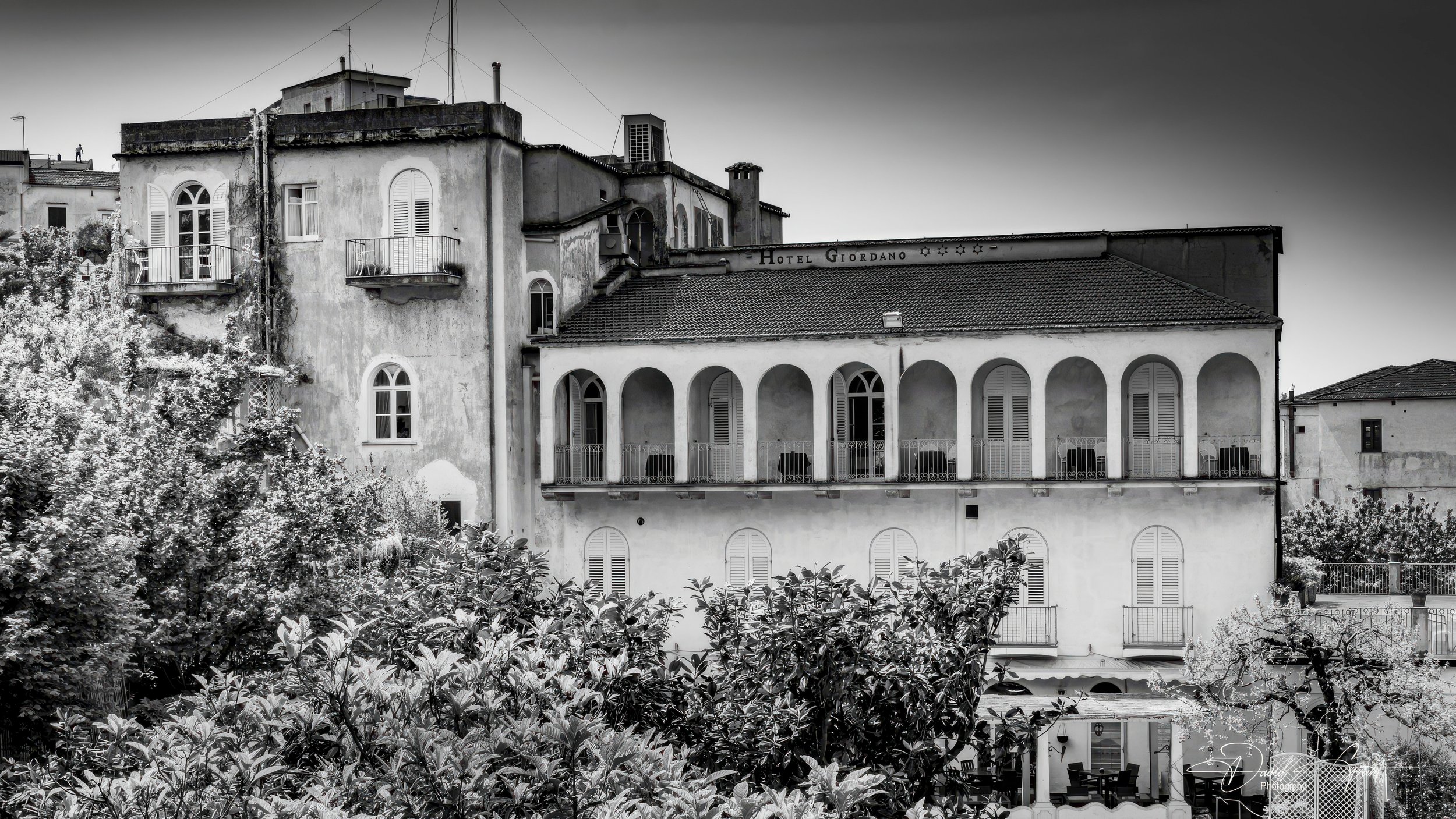 Black and white photo of an old building with a sign that reads 'Hotel Giordano'. The building has arched windows and balconies, surrounded by trees and foliage, with a cloudy sky overhead.