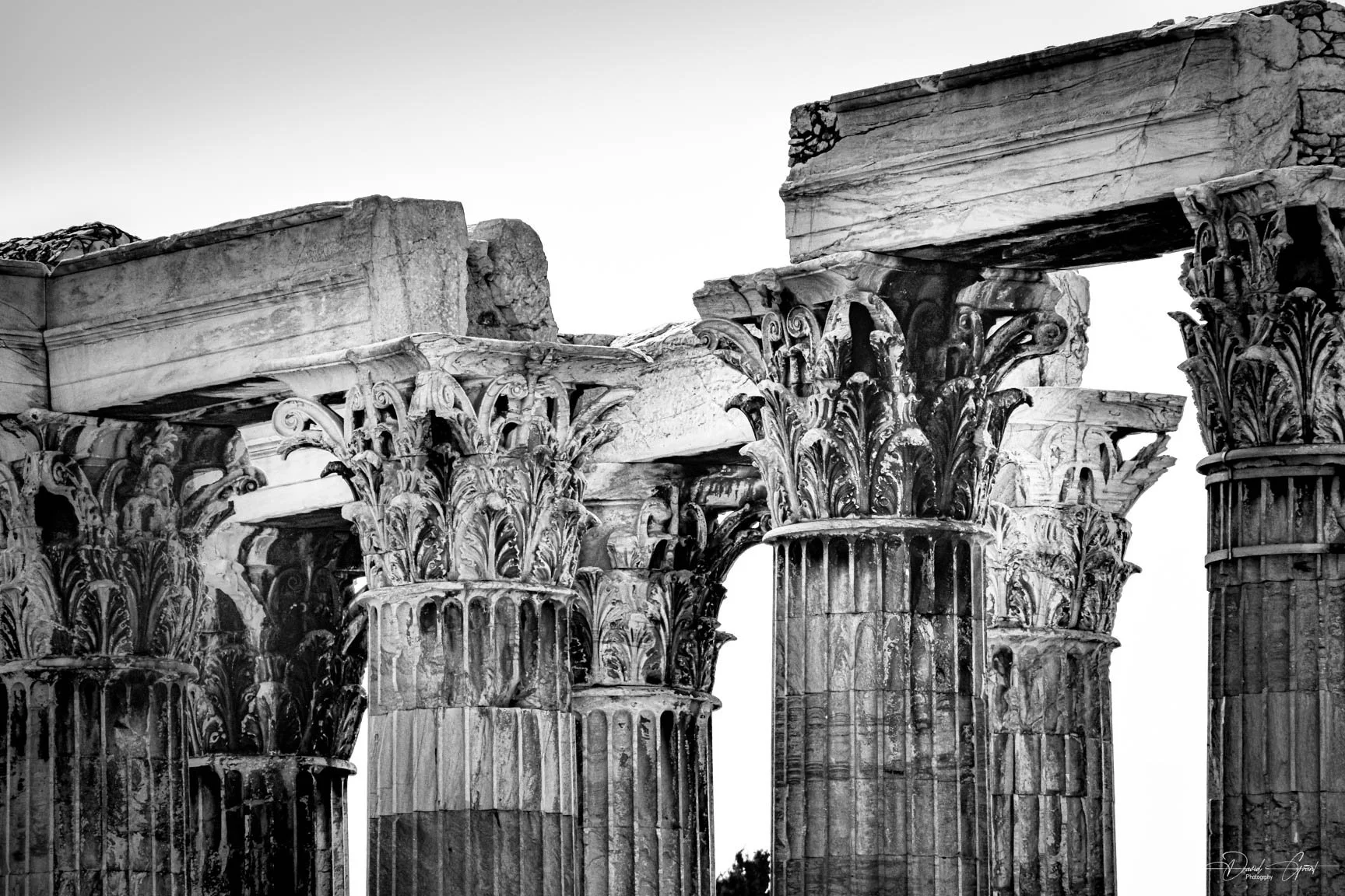 Black and white photo of ancient Greek temple ruins with ornate Corinthian columns and damaged stone entablature.