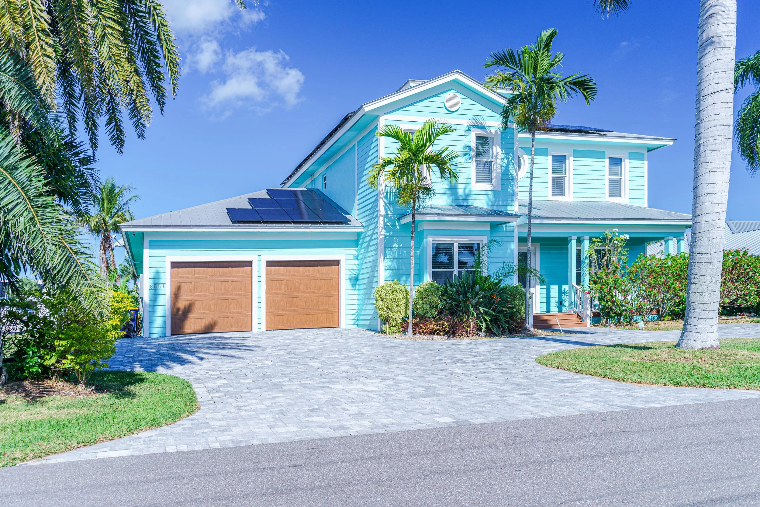 A bright blue two-story house with solar panels on the roof, surrounded by palm trees and lush greenery, under a clear blue sky.