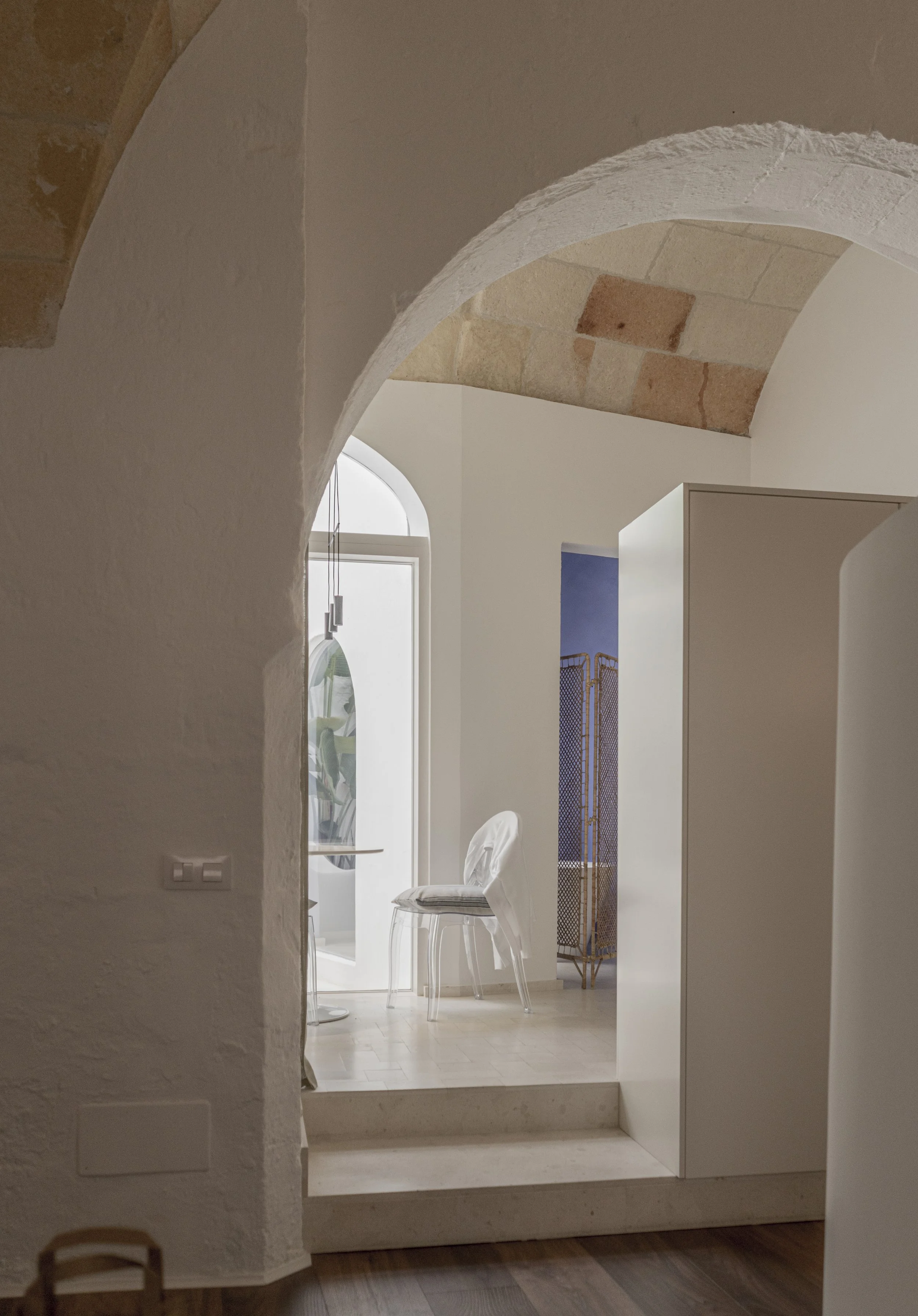 Interior view of a room with stone archways, a white chair, a table, an umbrella, and a decorative folding screen.