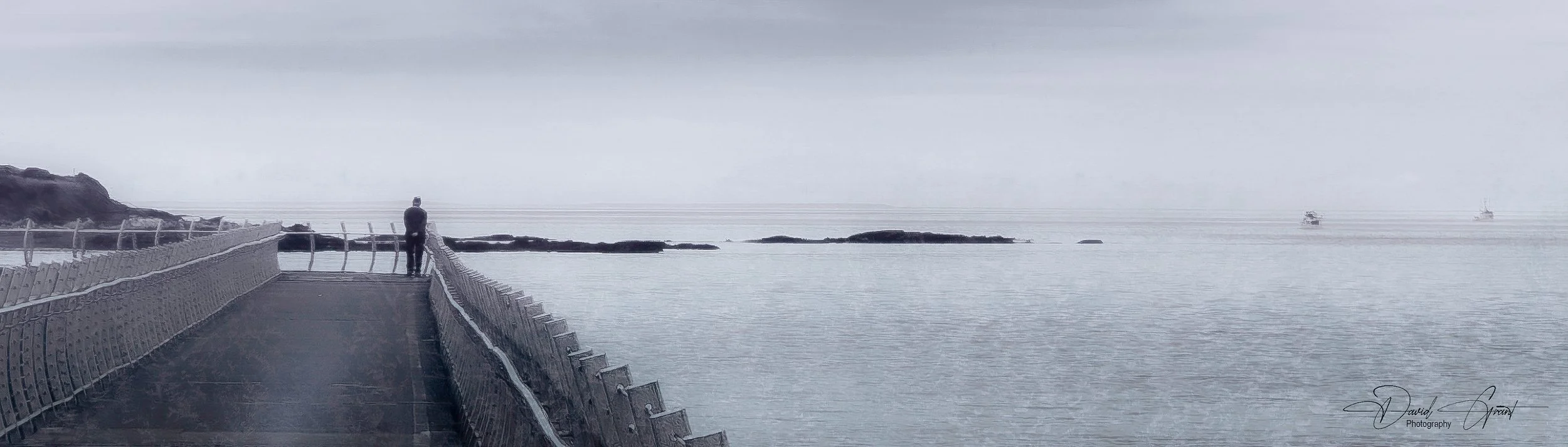 A person standing on a curved pier looking out over a calm sea on a cloudy day, with two ships visible in the distance.