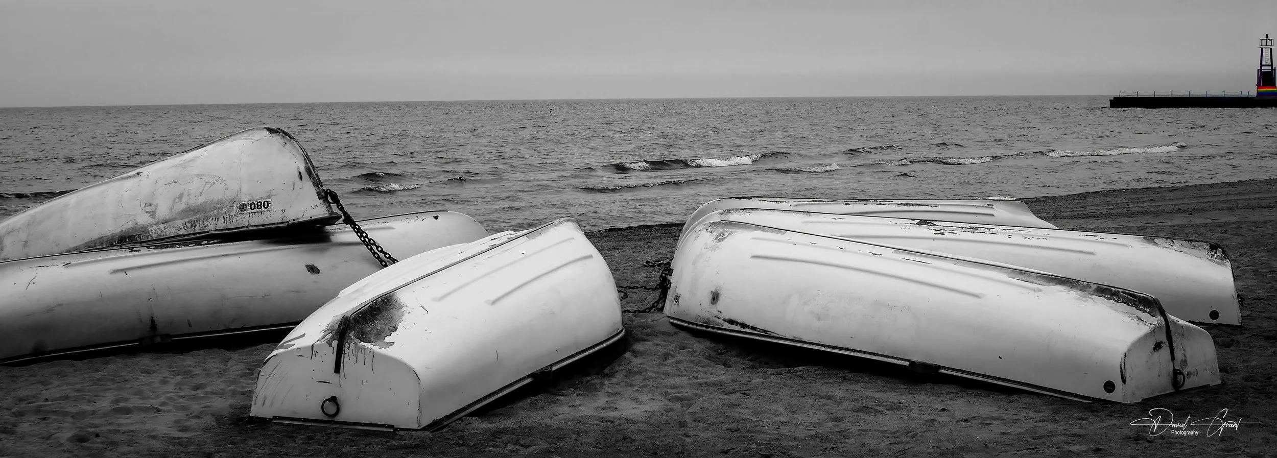 Five white rowboats upside down on the sandy beach with the ocean in the background and a small lighthouse on the horizon.