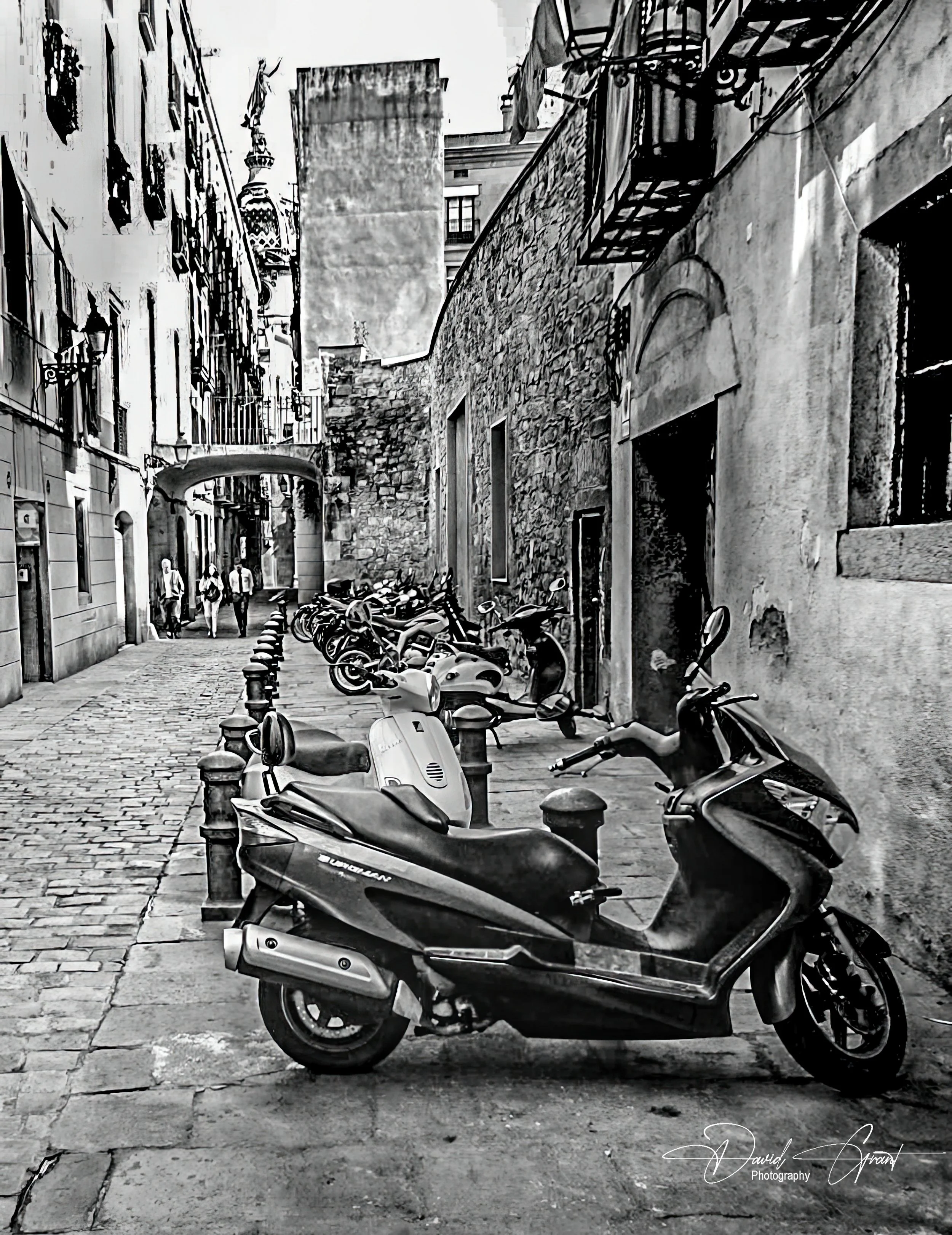 Black and white photo of a narrow cobblestone street with parked scooters and motorcycles along a stone and plaster building, with pedestrians walking in the background.