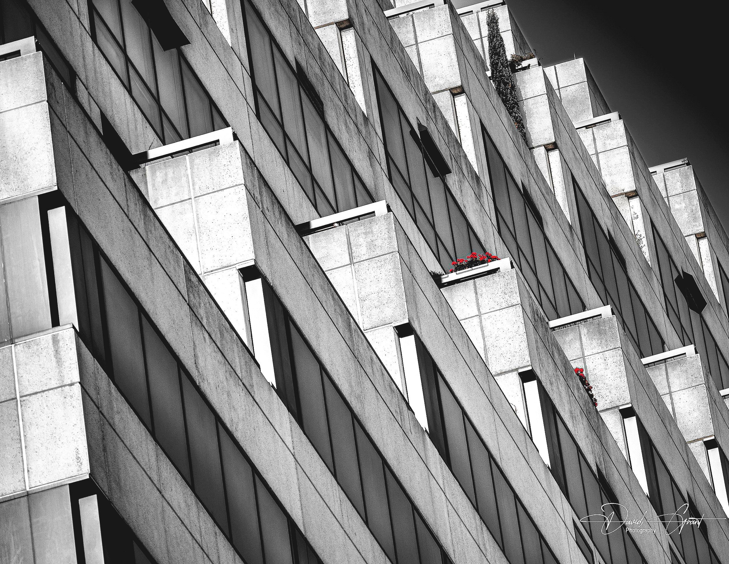 Close-up of a modern building with geometric concrete and glass facade, some balconies with red flowers.