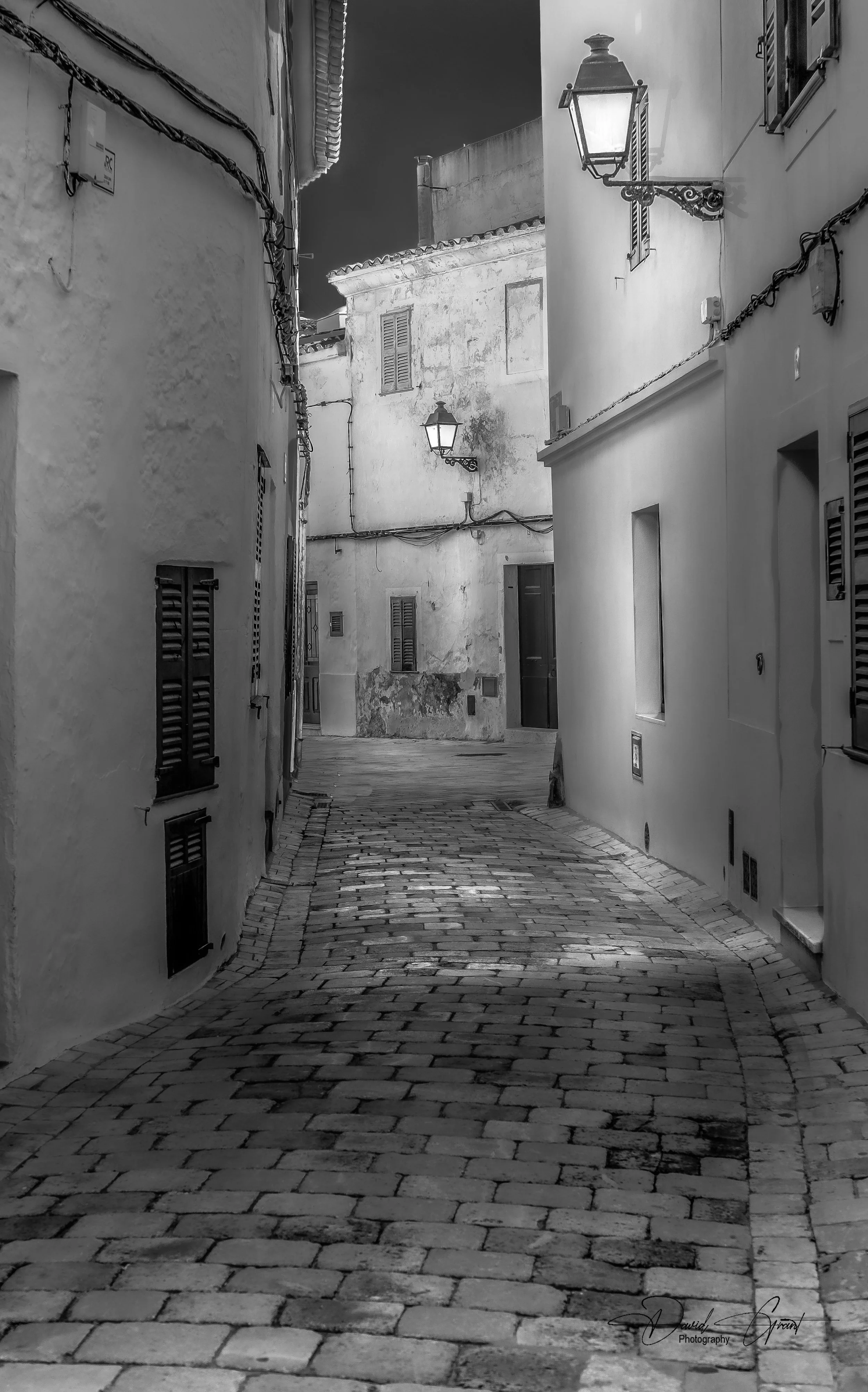 A narrow cobblestone street in a European village at night, illuminated by street lamps, with white plaster buildings and window shutters on either side.