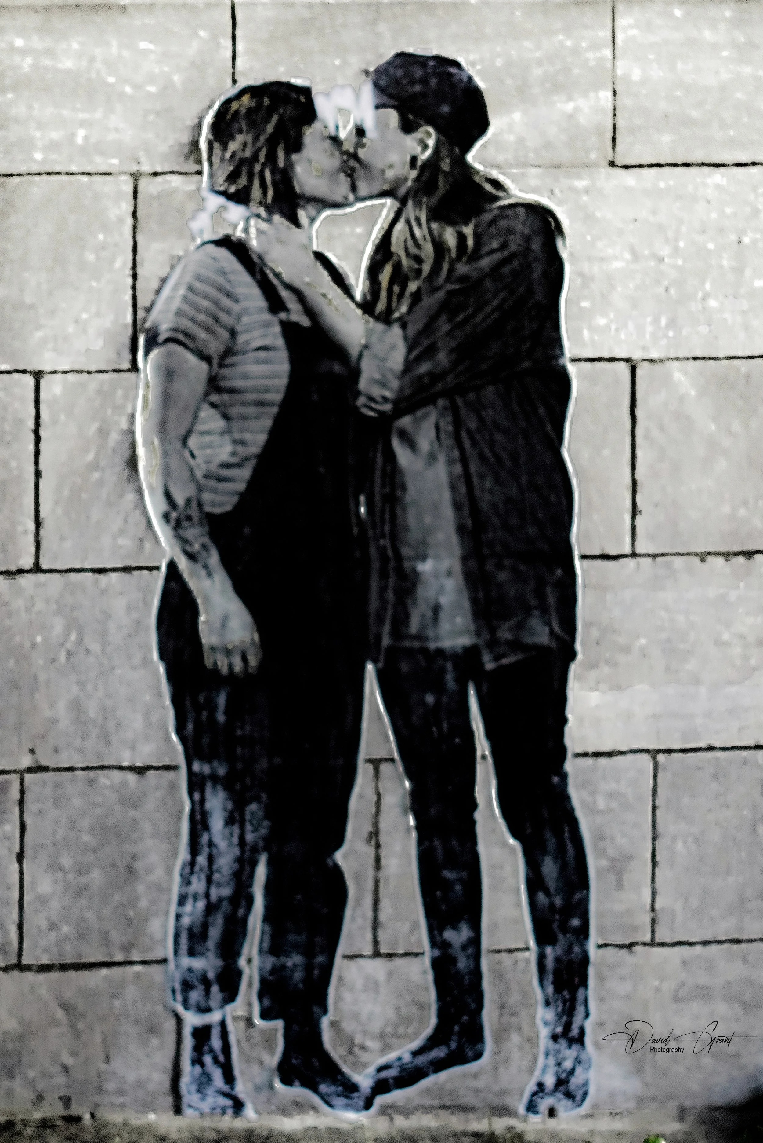 Street art of two women kissing, one with short hair, wearing a striped shirt and tattoos, the other with long hair and glasses, wearing dark clothing, on a brick wall background.